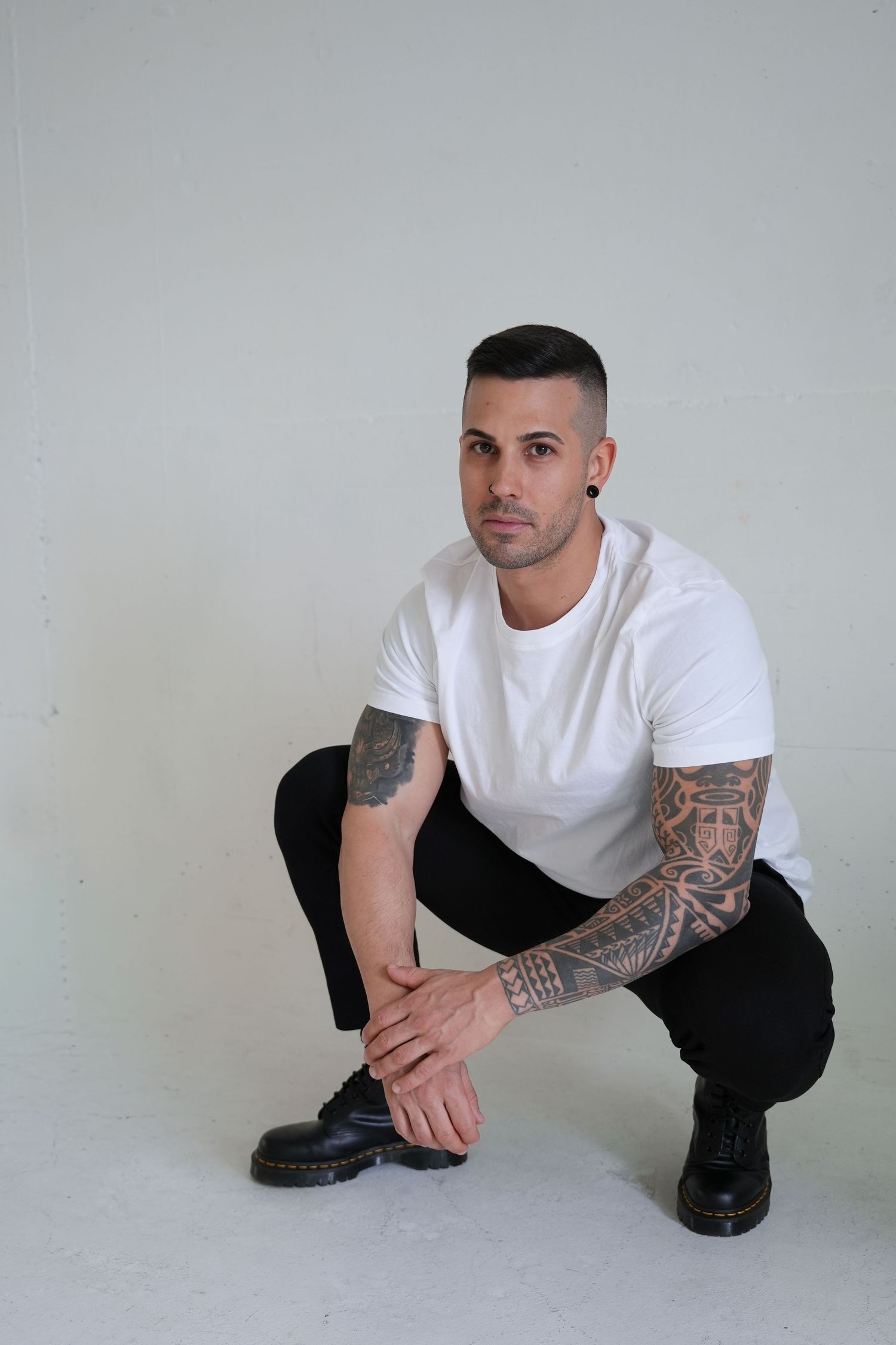 Tattooed man in white t-shirt and black jeans crouching against white wall wearing black boots.
