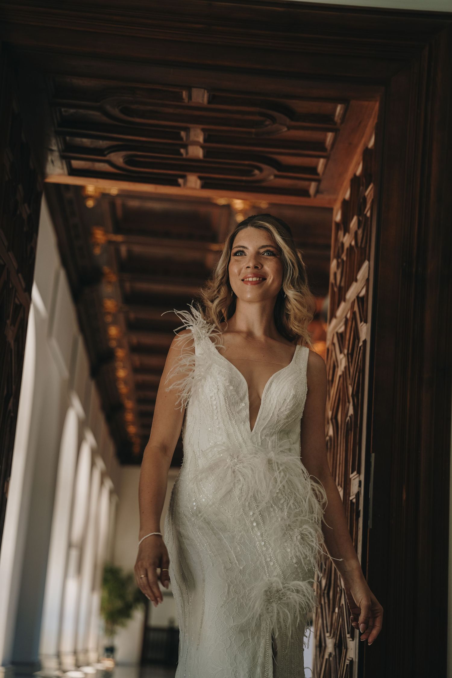 Bride in a white feathered lace wedding gown smiling in an elegant corridor with wooden ceiling.