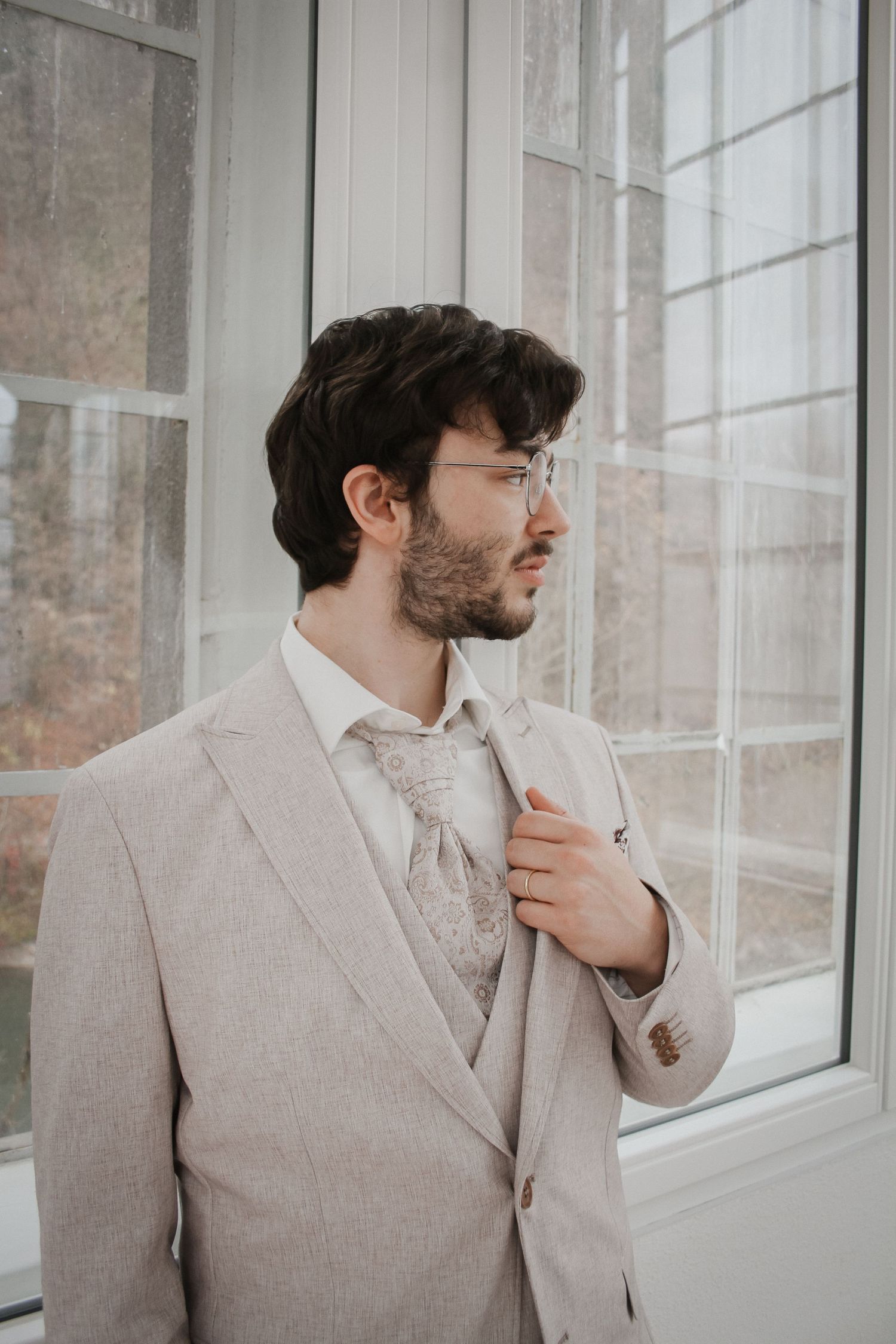 A well-dressed groom in a beige suit and floral tie adjusts his lapel while gazing out a window on his wedding day.