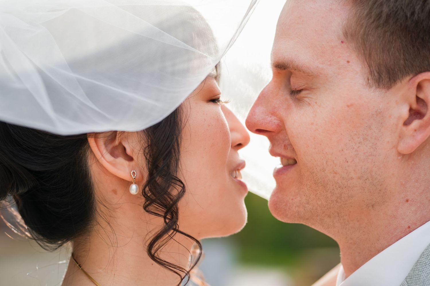 A bride and groom share a romantic almost-kiss moment, with wedding veil draped over them both.