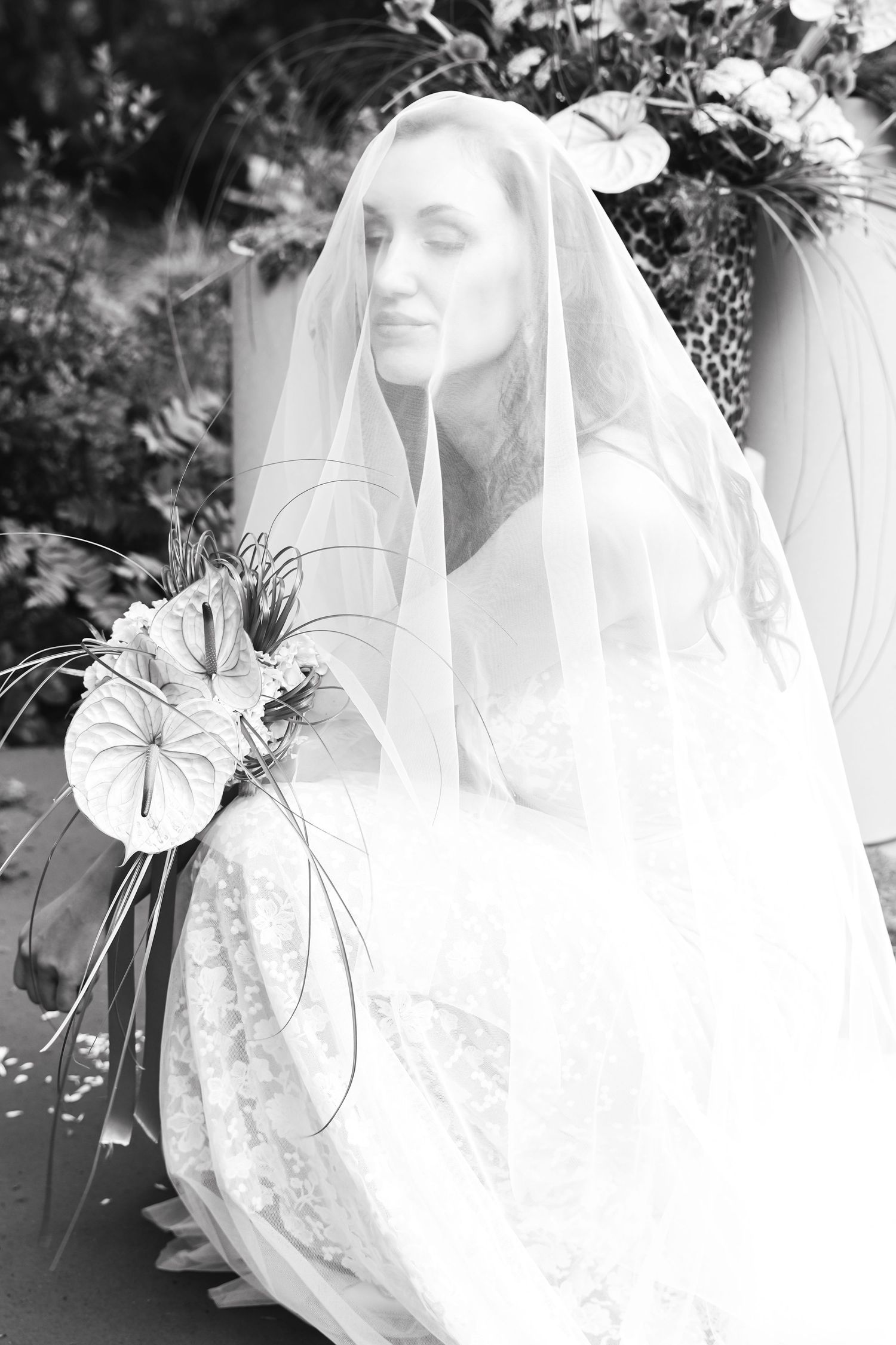Black and white portrait of a bride wearing a lace gown and veil, holding a bouquet, with floral arrangements in the background.