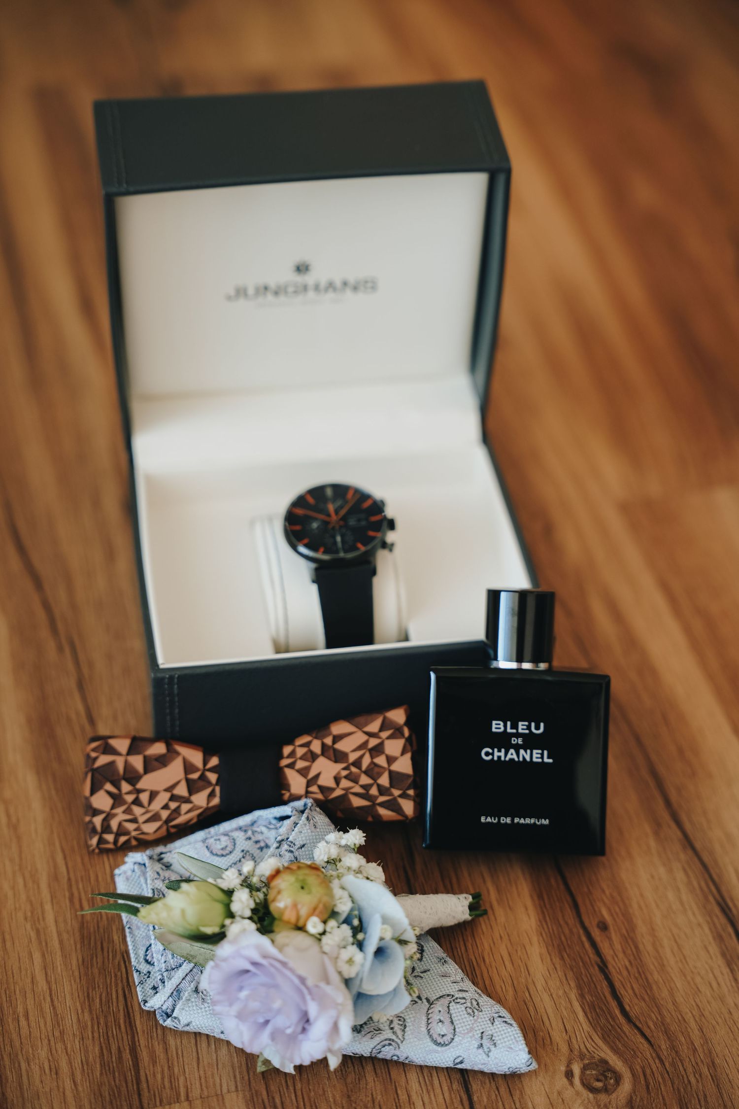Junghans watch in open box beside Bleu de Chanel perfume, wooden bow tie and floral boutonniere on a wood table.