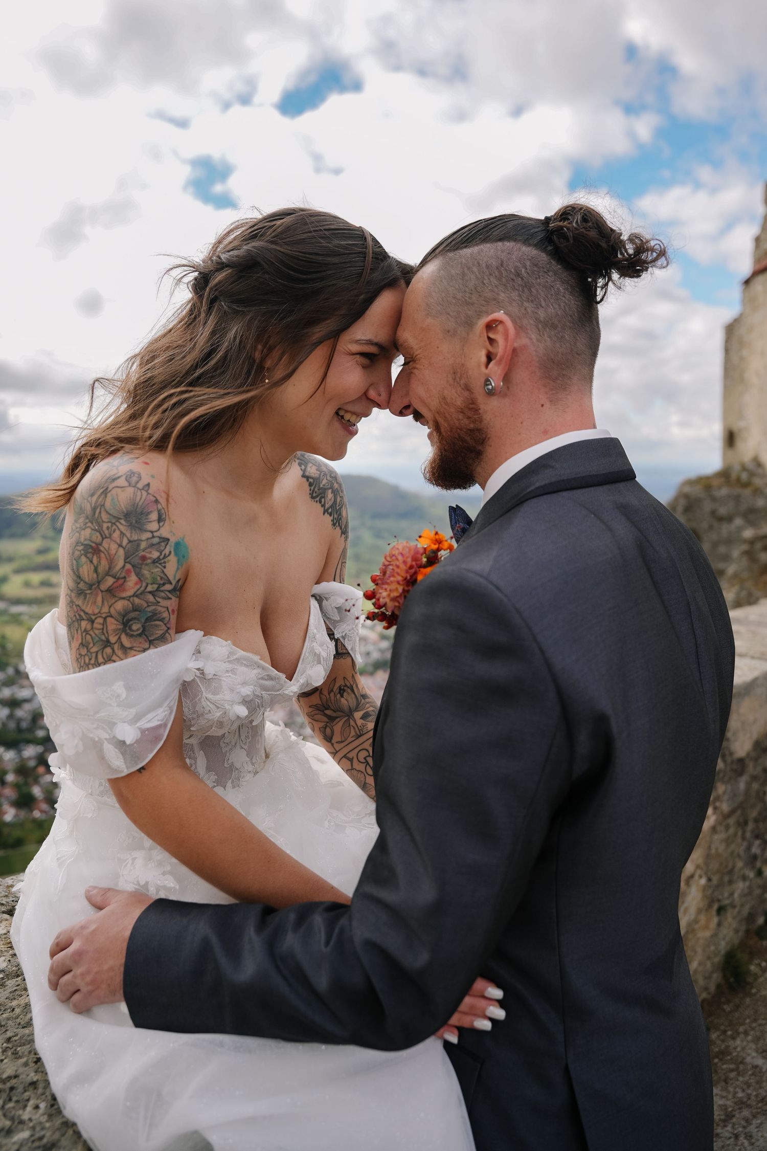 Smiling bride with floral tattoos and groom with a man bun touch foreheads on a rocky hilltop wedding day with cloudy sky and valley views.