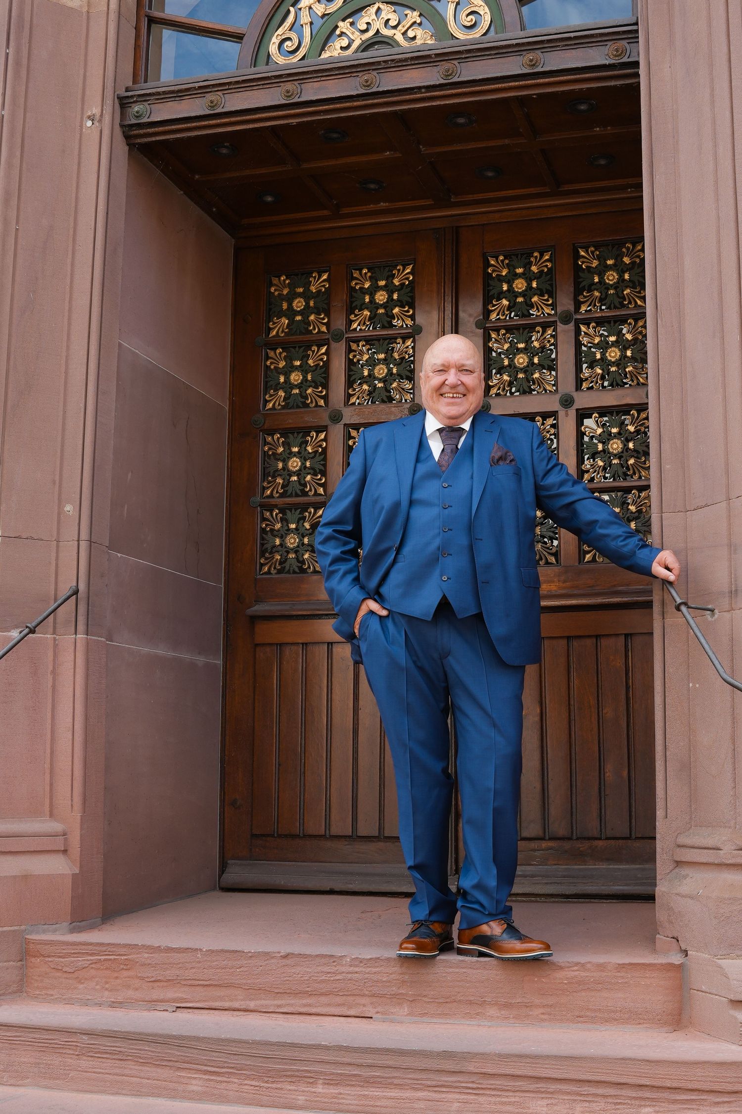 Man in blue three-piece suit standing at ornate wooden doors of elegant building entrance.