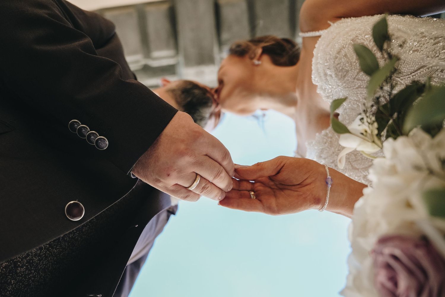 Bride and groom exchanging wedding rings during ceremony, close-up of hands with bouquet of white flowers.