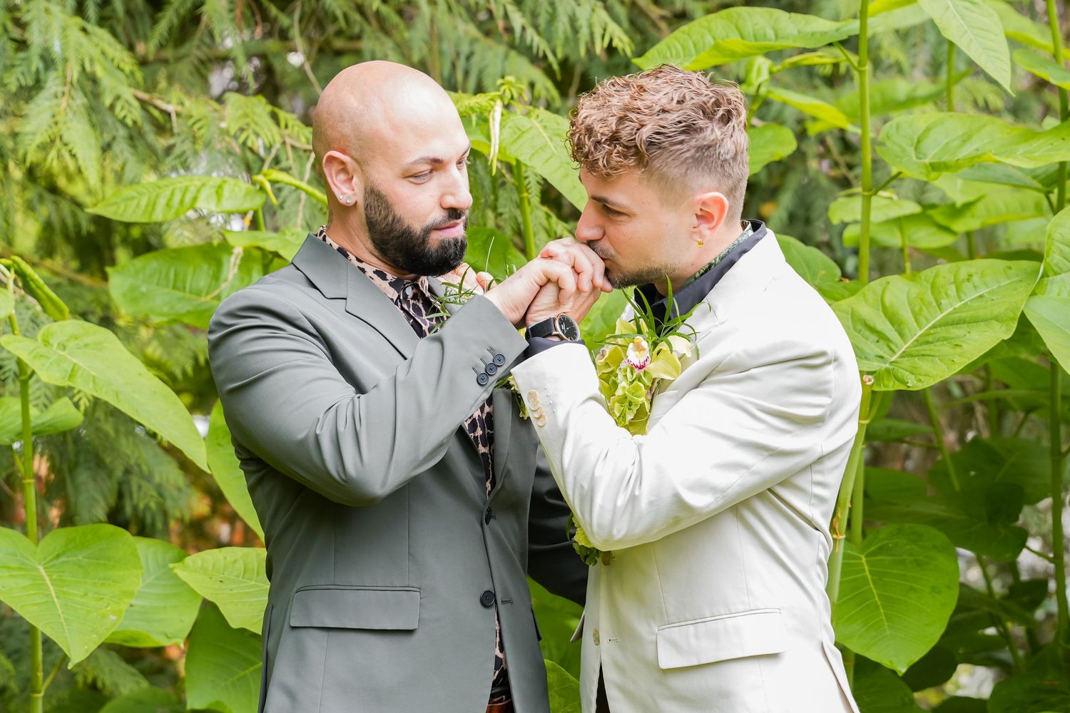 Two grooms in suits share a tender hand-kissing moment outdoors surrounded by lush green foliage.