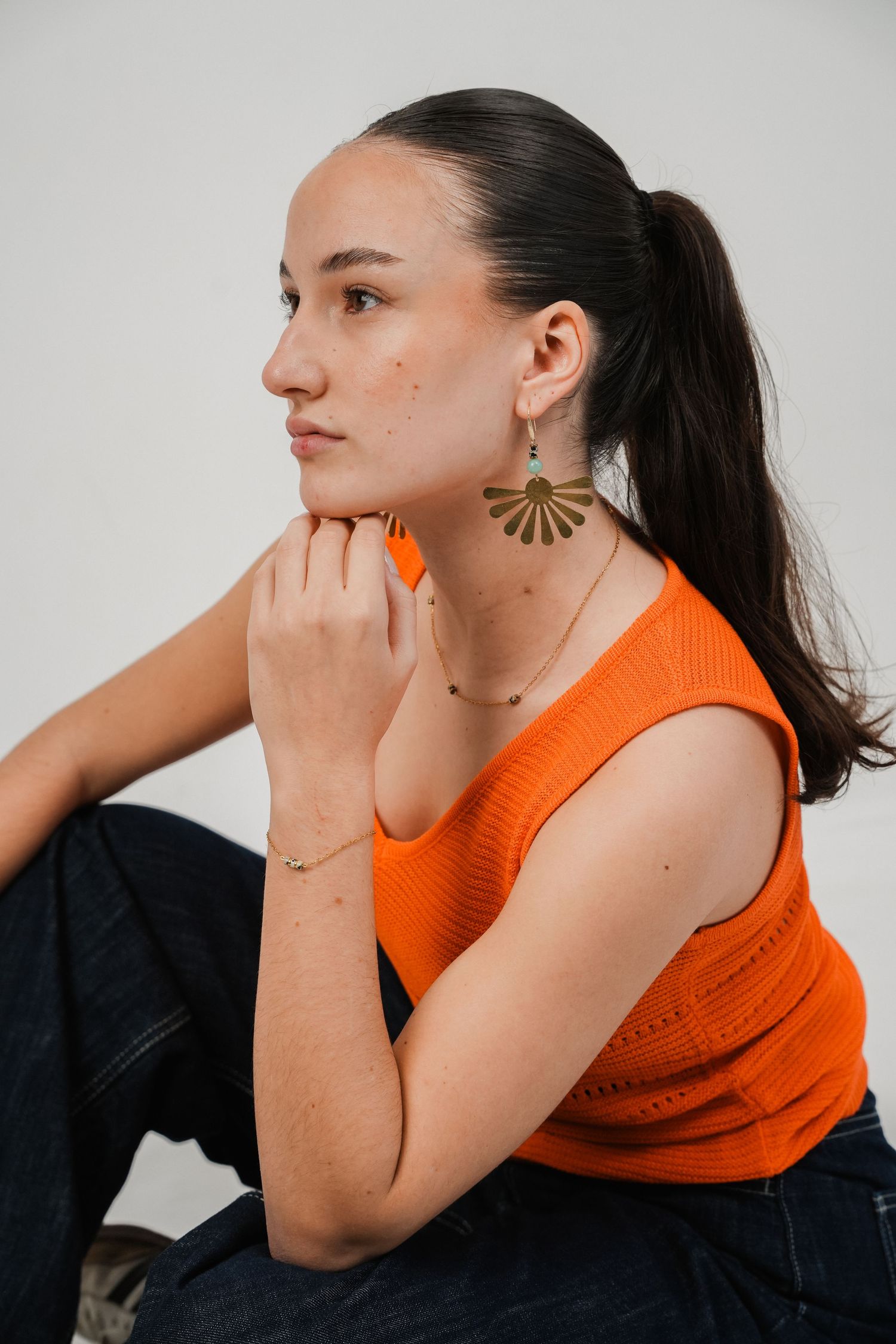 Woman in orange sleeveless top wearing gold fan earrings and delicate necklace, sitting pensively against white background.