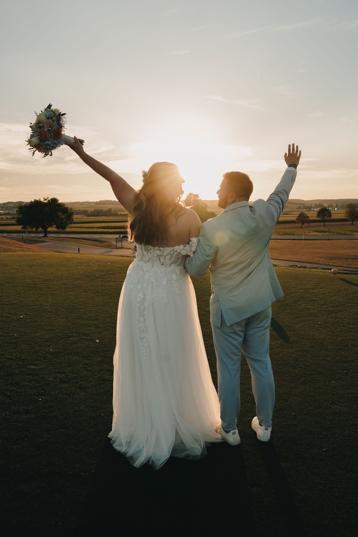 Bride and groom celebrating at sunset, arms raised, overlooking a scenic green landscape.