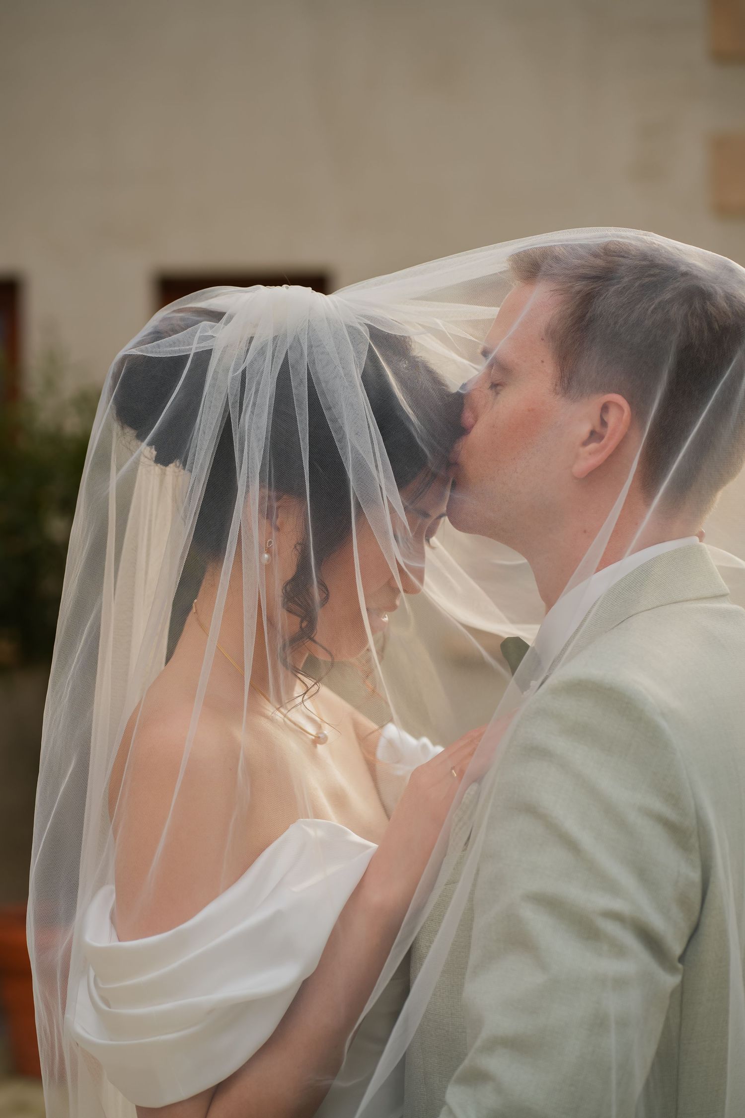 Bride and groom sharing a romantic kiss under a flowing white wedding veil outdoors.