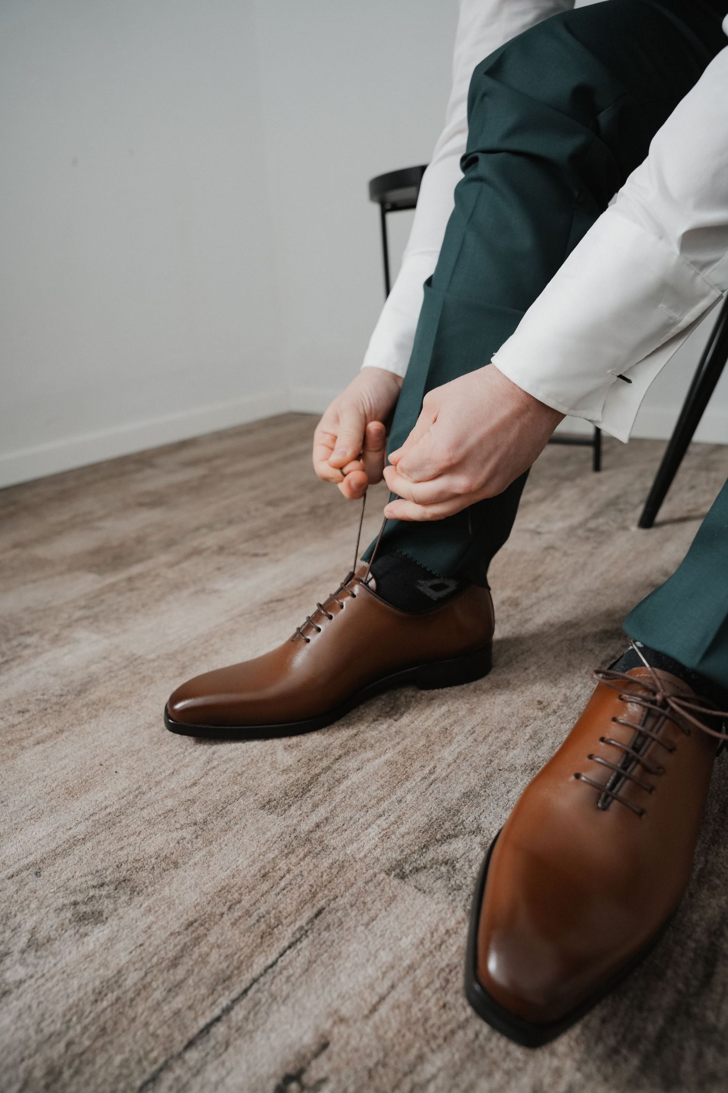 Man in green trousers tying brown leather Oxford dress shoes while seated, elegant formal footwear on wooden floor.