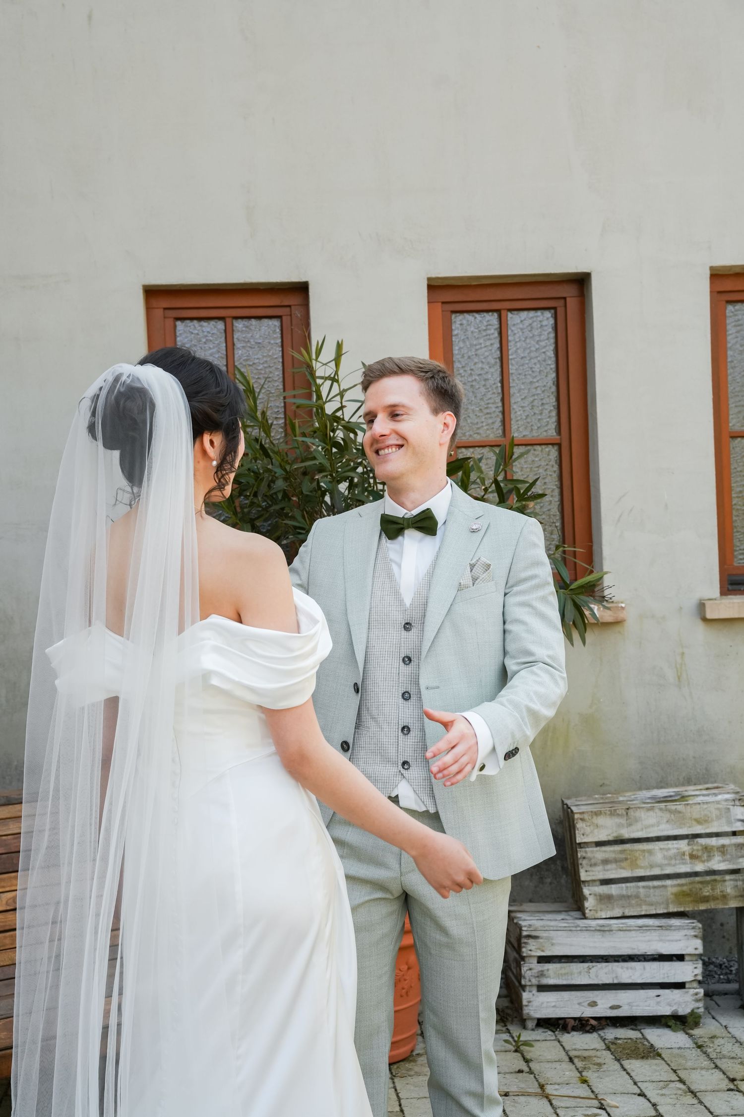 Smiling groom in light gray suit with bow tie holds bride's hand outdoors during wedding ceremony.