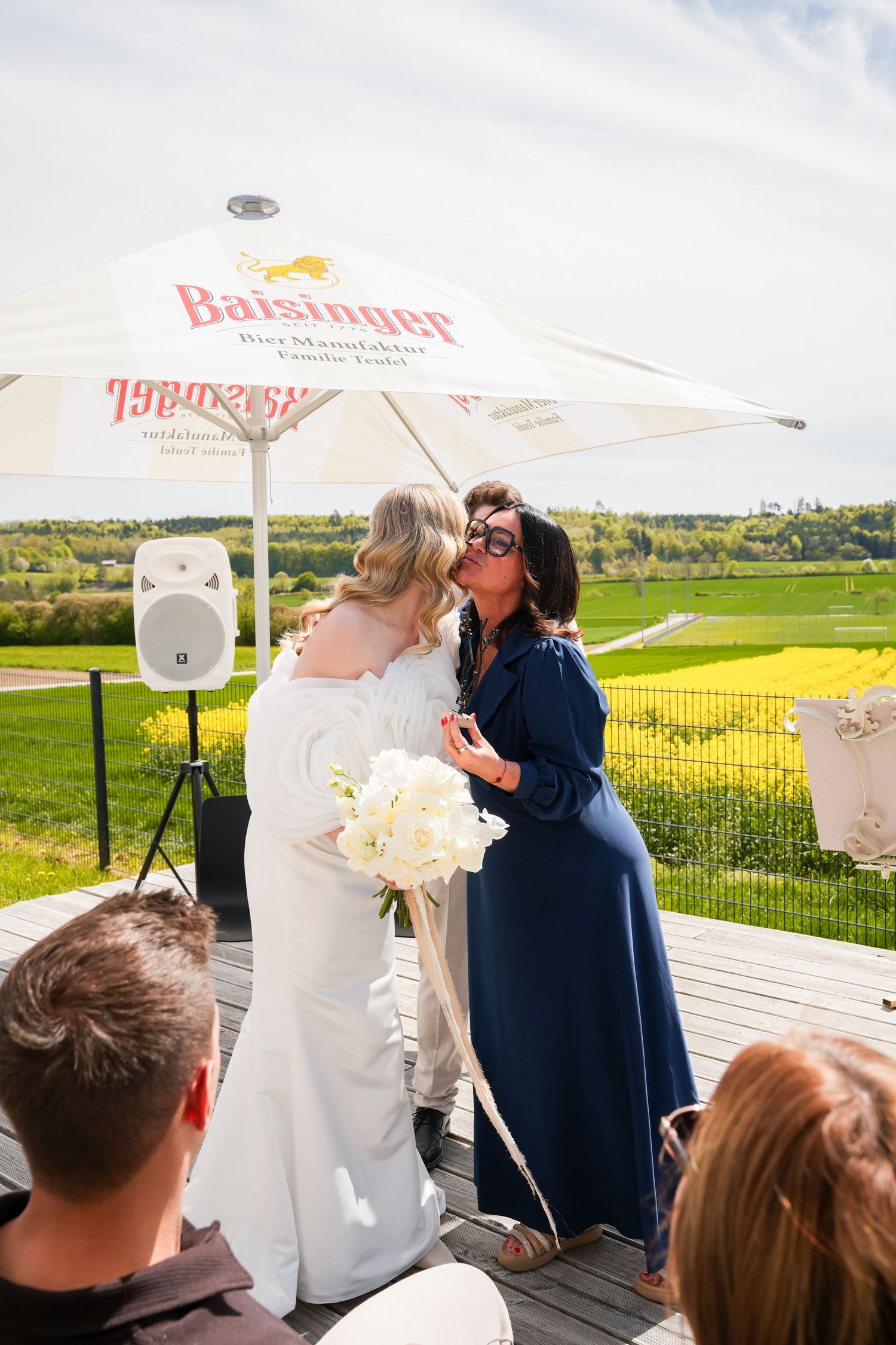 Bride in white gown embracing guest in blue dress outdoors at wedding venue with green fields backdrop.