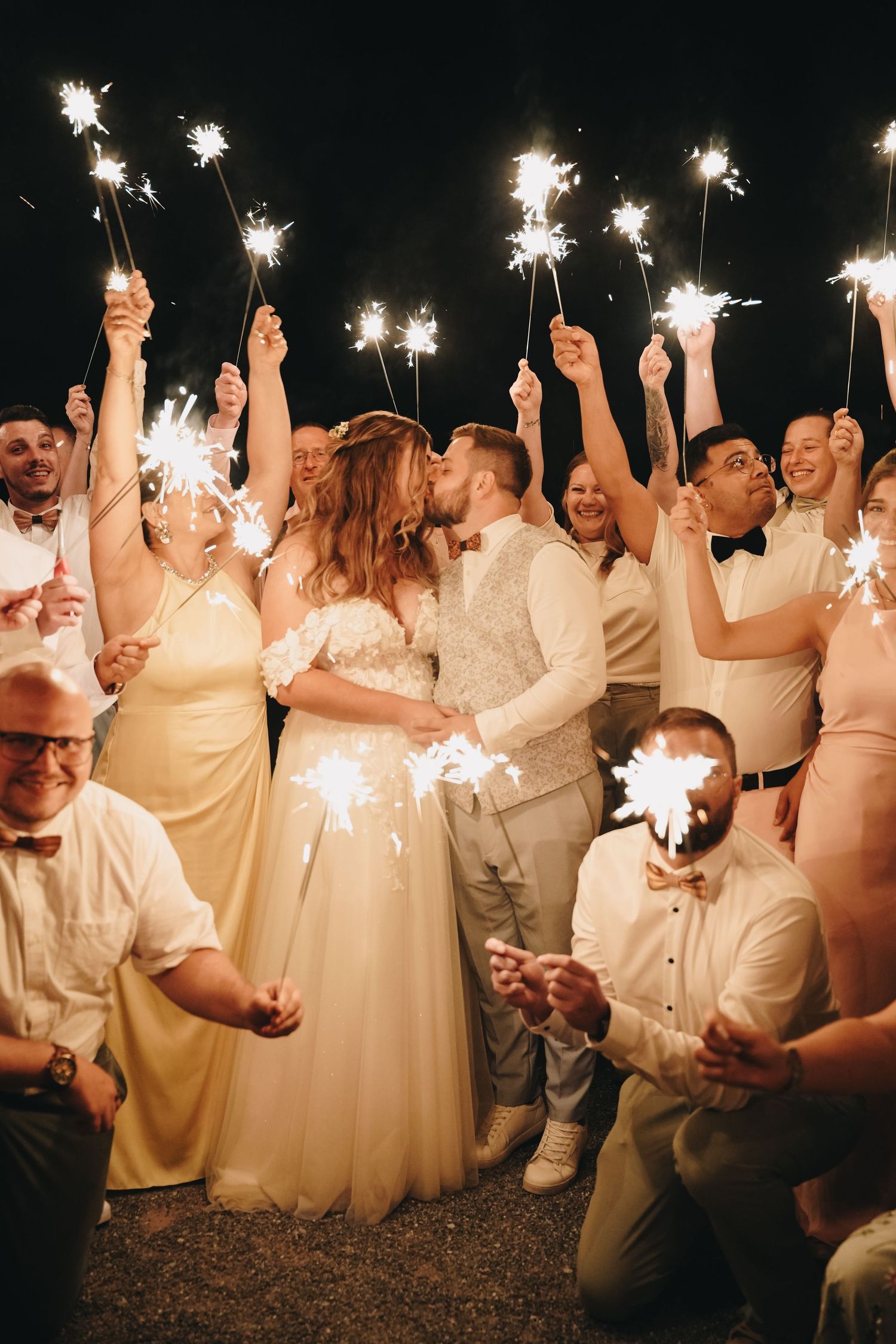 Bride and groom kiss at night while wedding guests in formal attire hold glowing sparklers in a joyful circle around them.