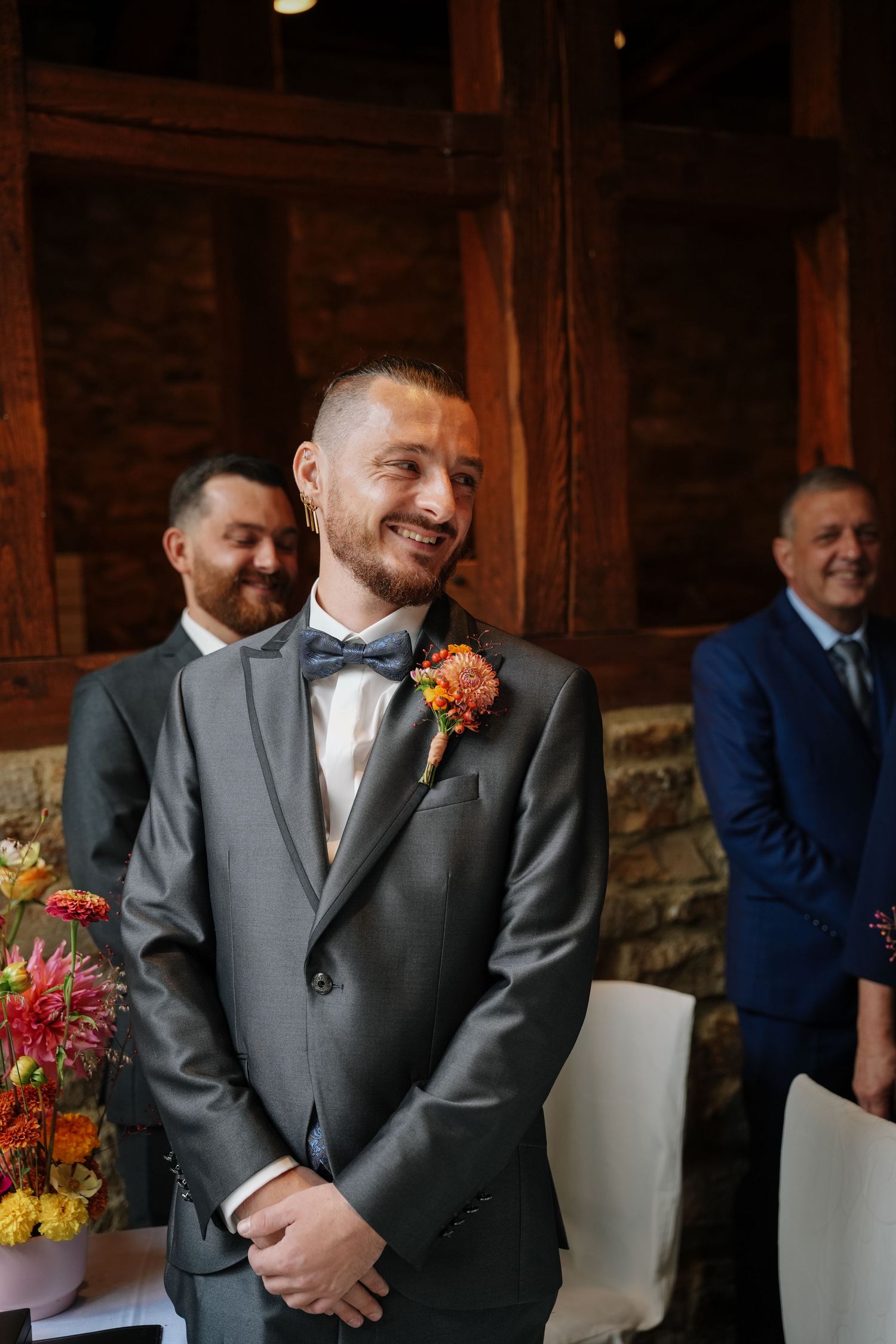 Happy groom with a beard and floral boutonniere smiles warmly while guests stand behind him in a timber-framed wedding venue.