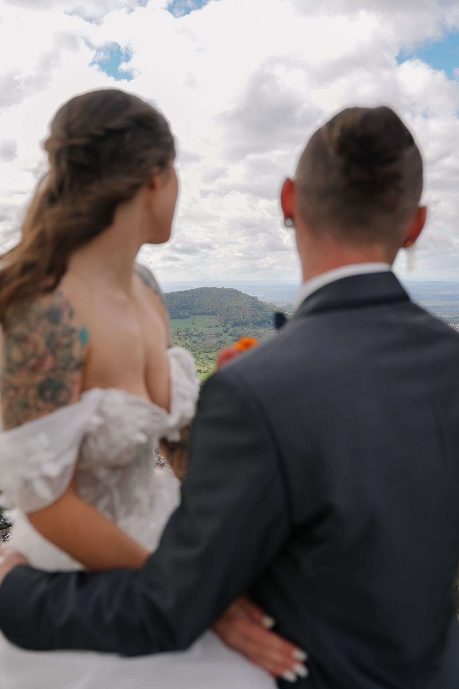 Bride in a white off-the-shoulder gown with a floral shoulder tattoo embraces a groom in a dark suit, gazing at a scenic hilltop view.