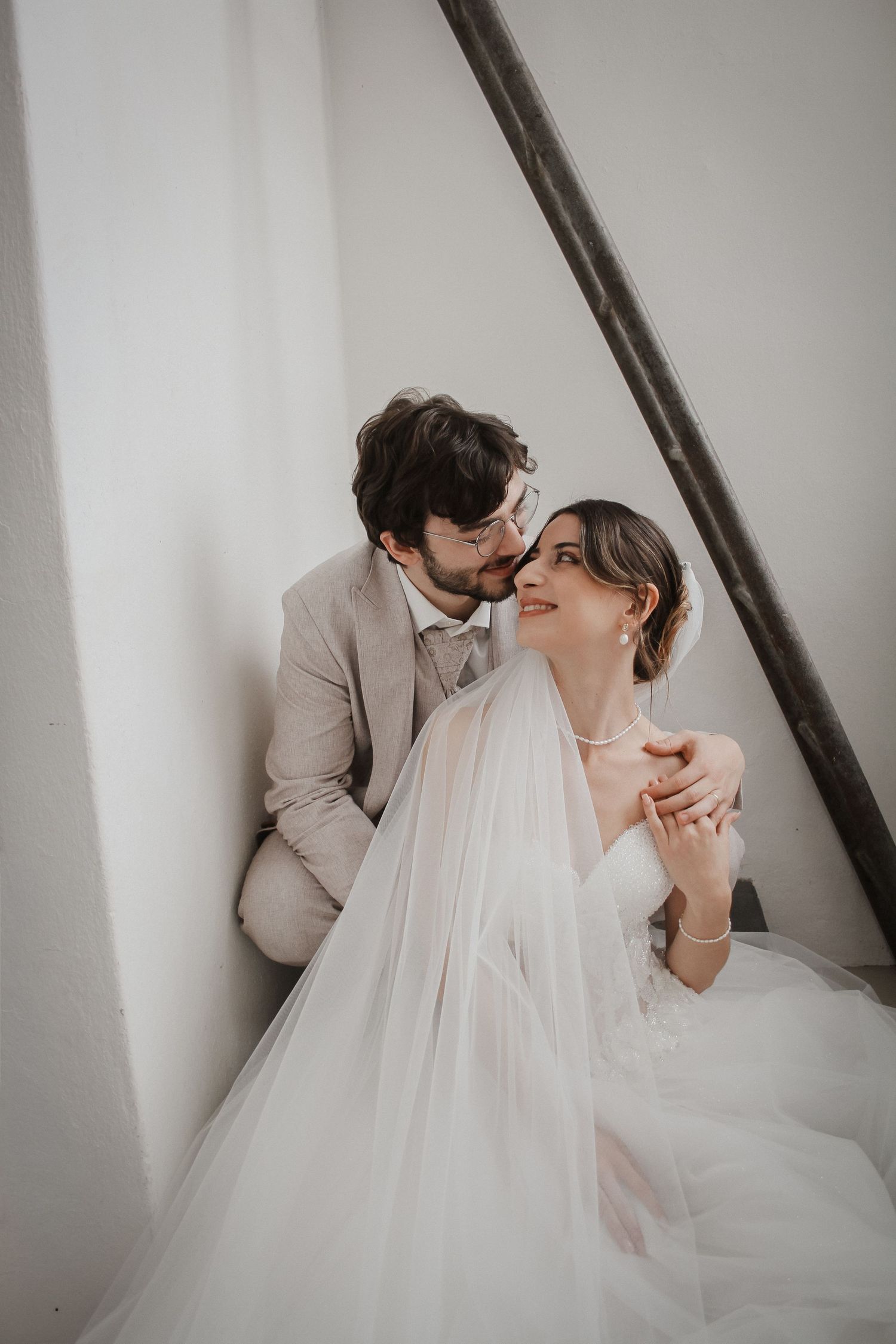 A bride in a white gown and veil sits with a groom in a beige suit as they share a tender moment together.