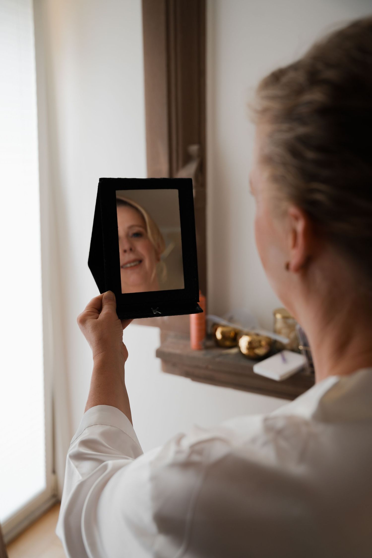 A woman in a white robe holds a small mirror, smiling at her reflection near a bright window.