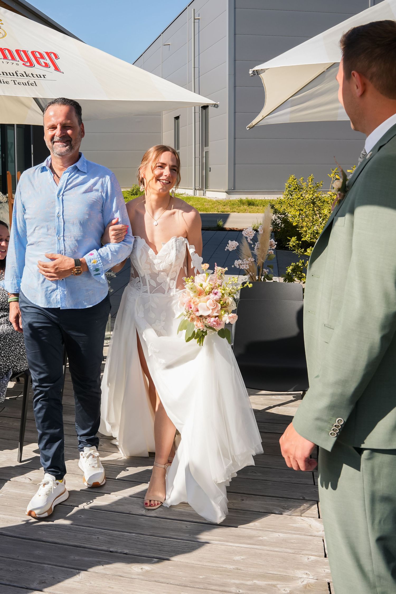 Smiling bride in white gown holding pink bouquet walks with man in blue shirt toward groom on sunny outdoor terrace.
