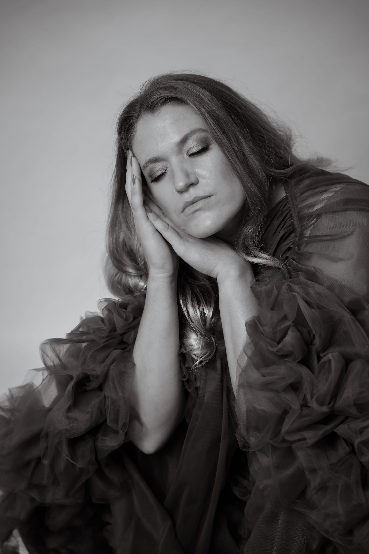 Black and white portrait of a woman in a ruffled tulle gown, eyes closed, hands framing her face.