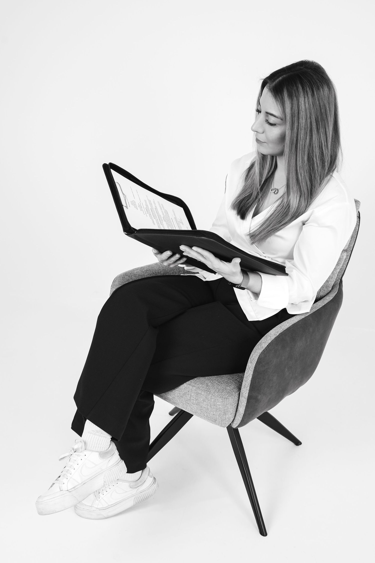 Black and white photo of a businesswoman sitting in a chair reading documents in a folder.
