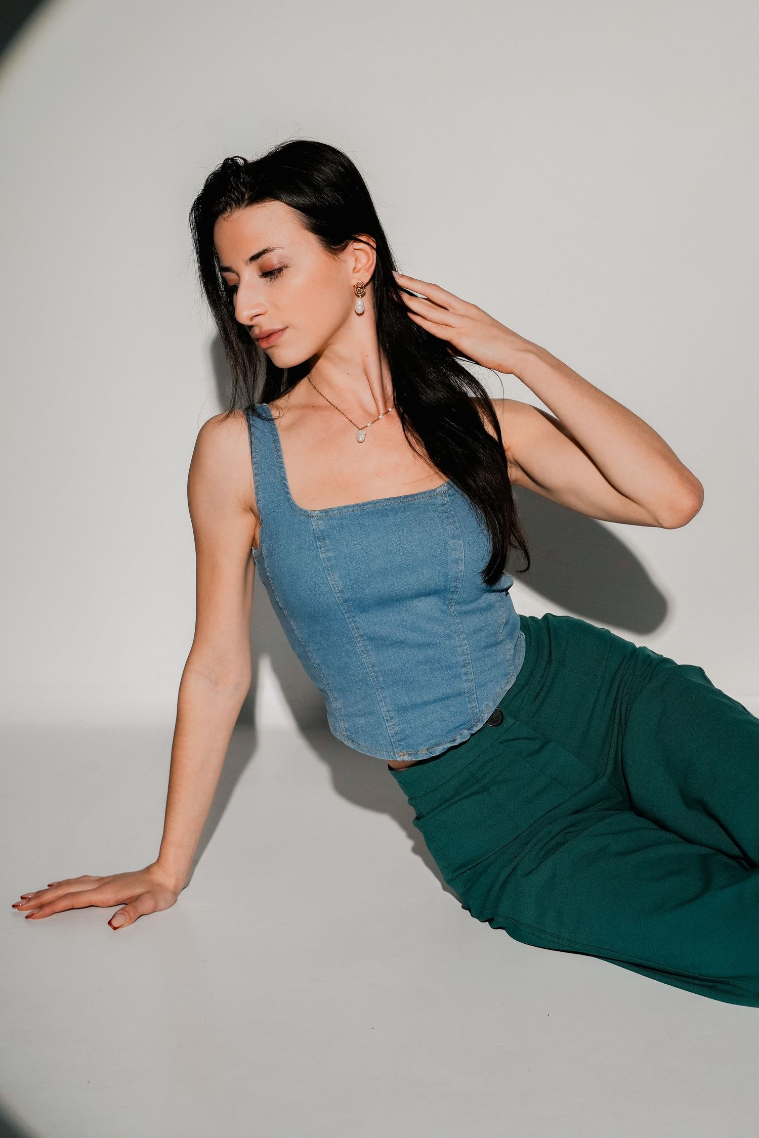 Woman in blue denim tank top and green trousers posing against white background with hand near neck.
