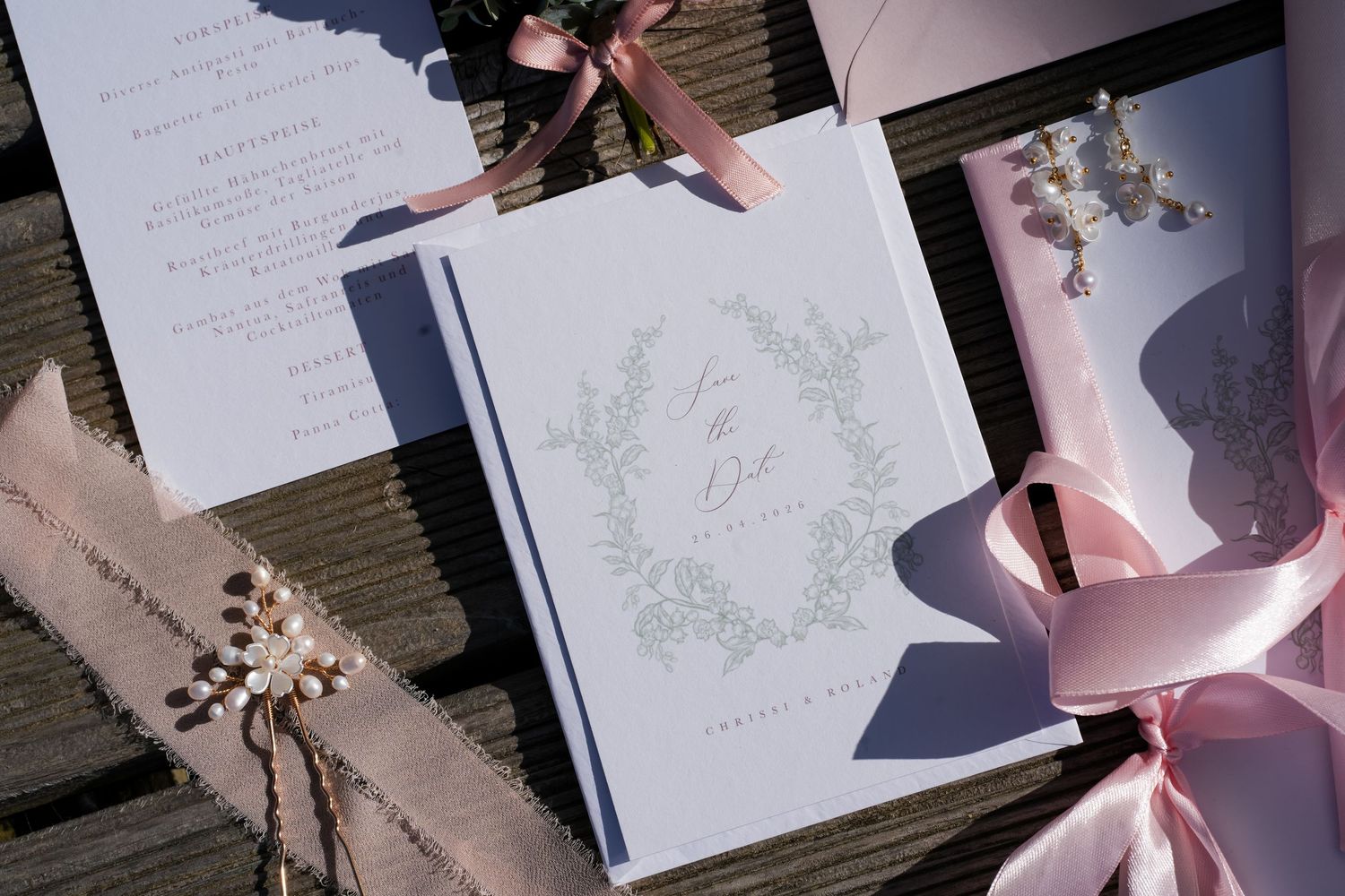Elegant wedding stationery suite with floral wreath design, pink ribbons, and pearl accents on rustic wood backdrop.
