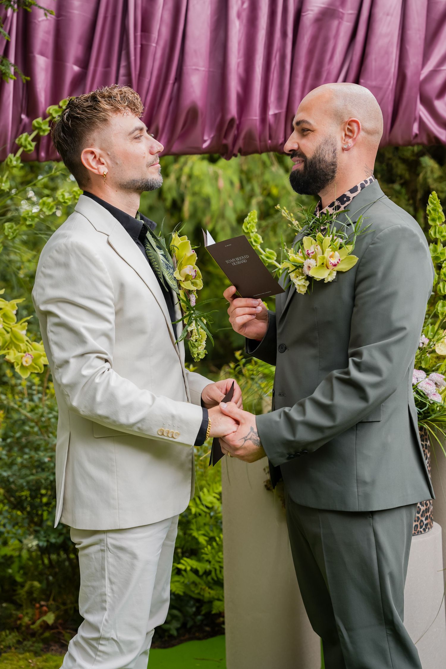 Two grooms exchange vows at an outdoor wedding ceremony surrounded by lush greenery and purple draping.