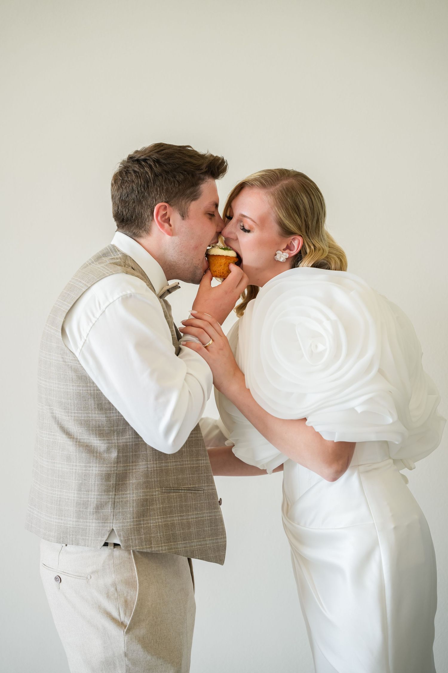 Bride and groom sharing an ice cream cone kiss in wedding attire, white backdrop.
