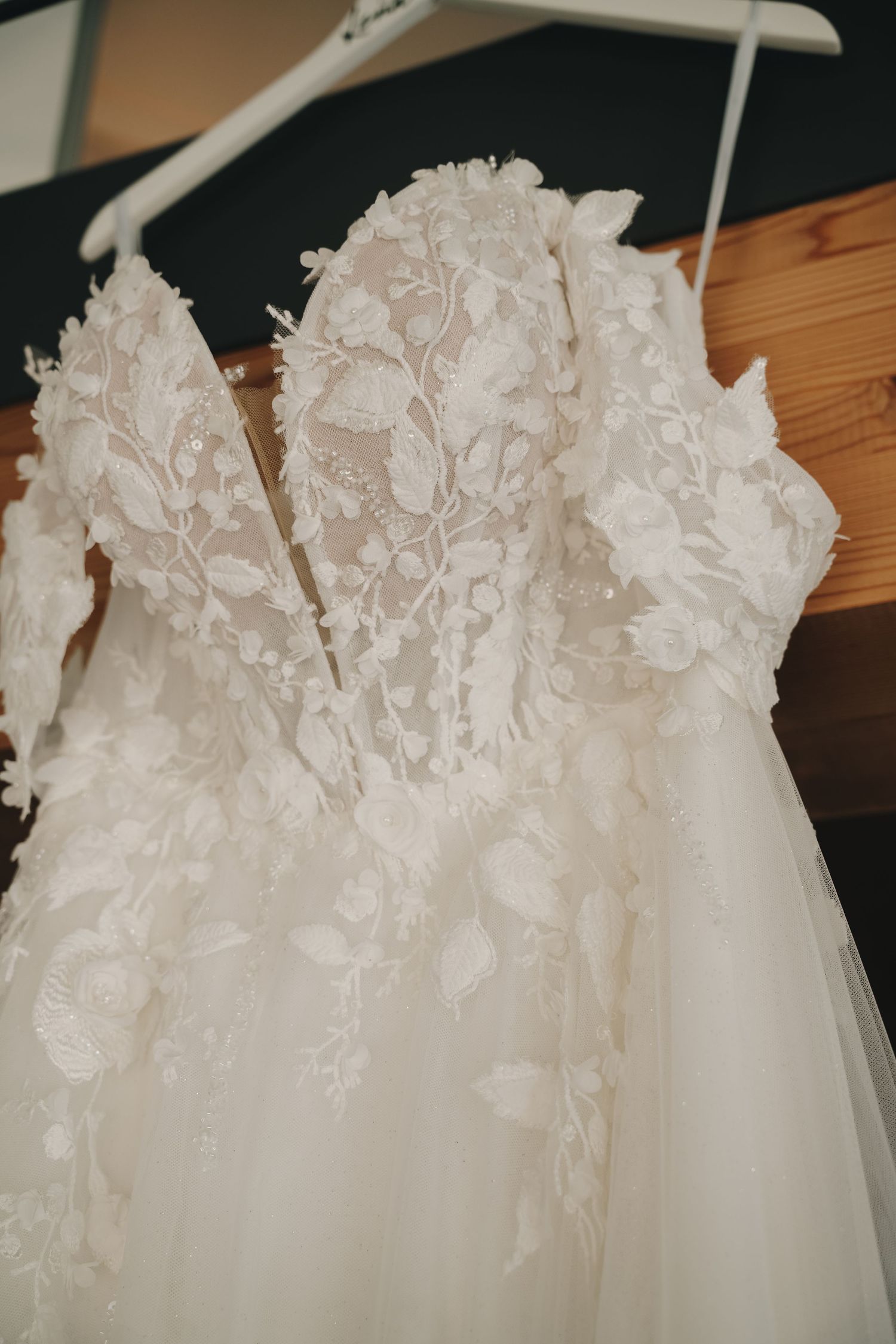 Elegant white wedding dress with 3D floral lace appliques and off-shoulder sleeves hanging on a white hanger.