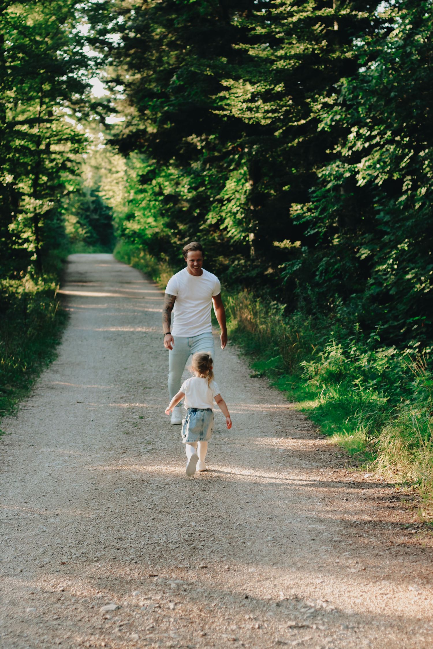 Father and young child running together on a scenic tree-lined dirt path in summer.