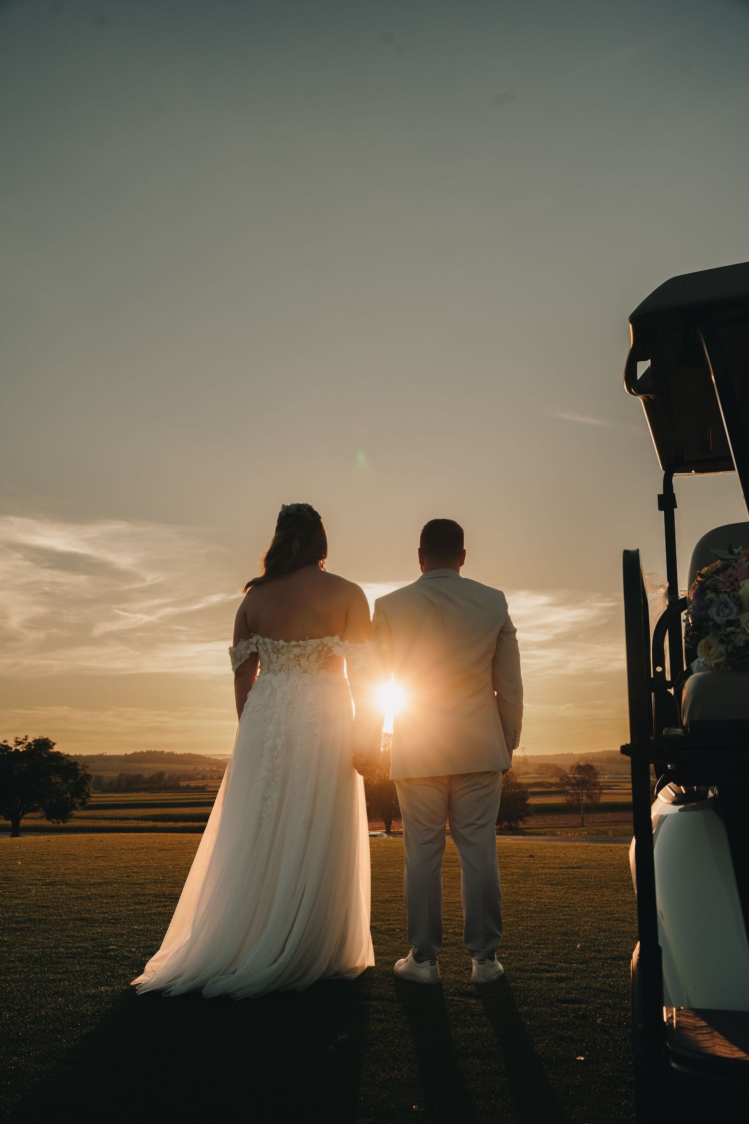 Bride and groom silhouetted against golden sunset on golf course, standing beside a golf cart.