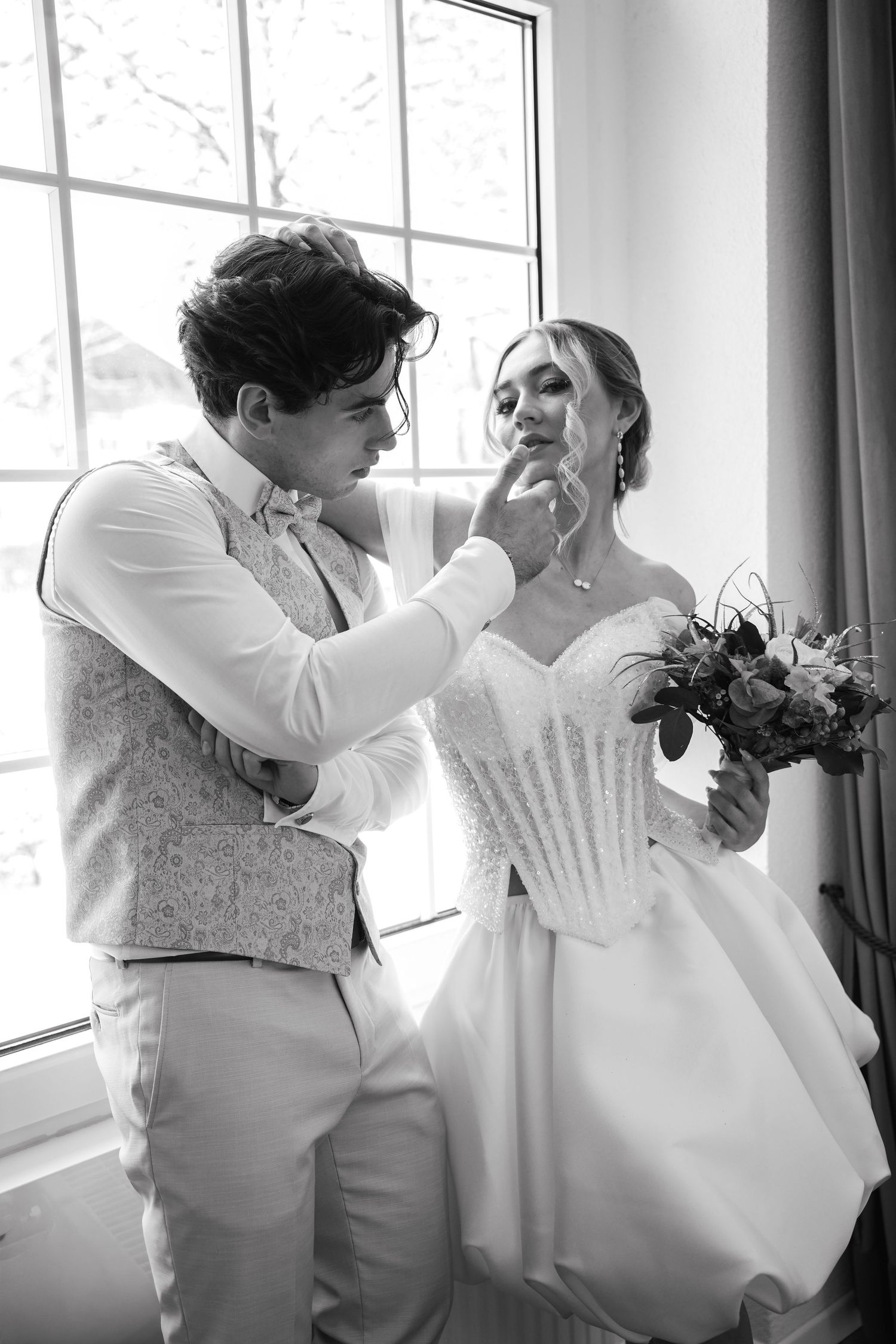 Black and white photo of a groom tenderly touching his bride's face near a window on their wedding day.