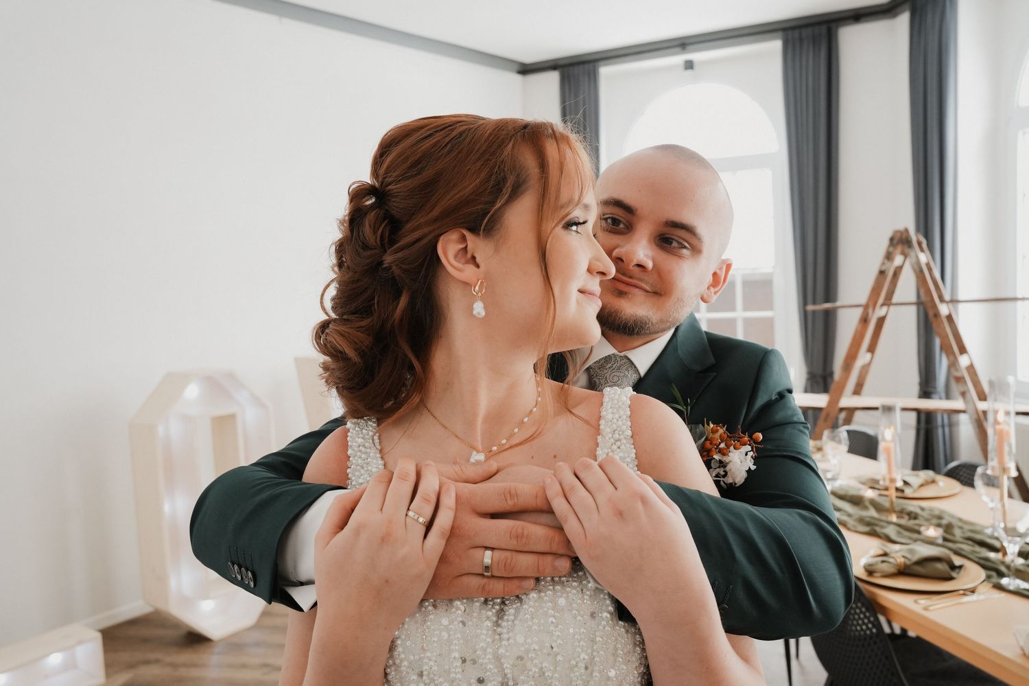 Bride and groom share a tender embrace, with groom in dark suit hugging bride in white lace dress indoors.