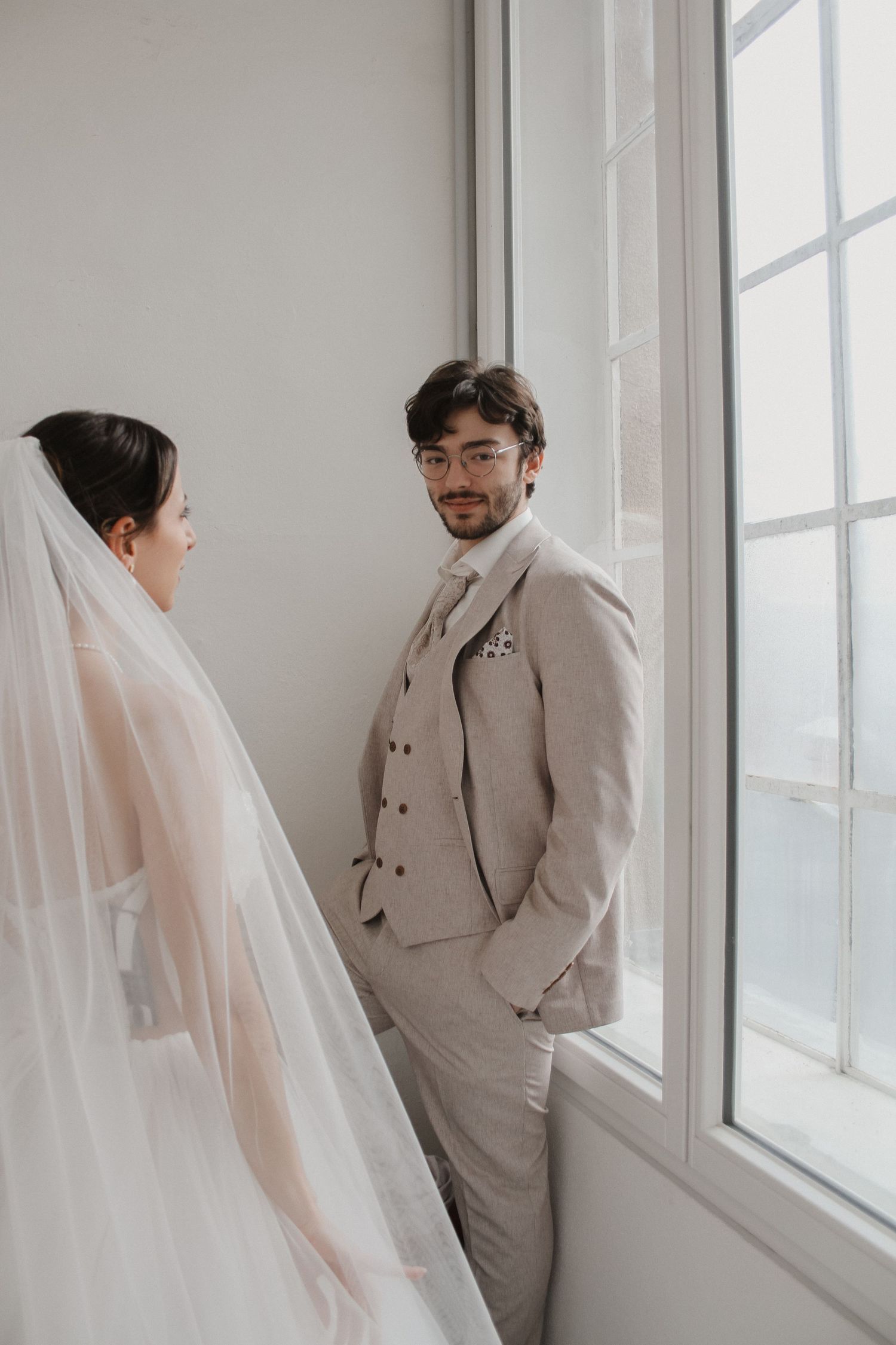 Groom in beige suit smiles at bride in wedding veil by large window in elegant indoor setting.