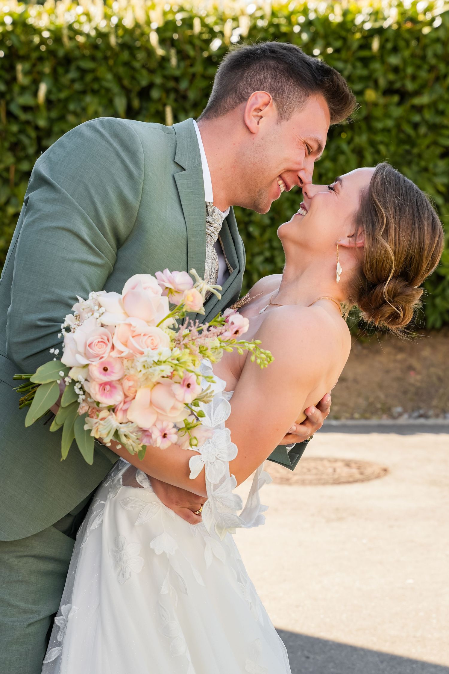 Bride and groom share a romantic moment, nearly kissing, as she holds a pink and white bouquet in a sunny outdoor setting.