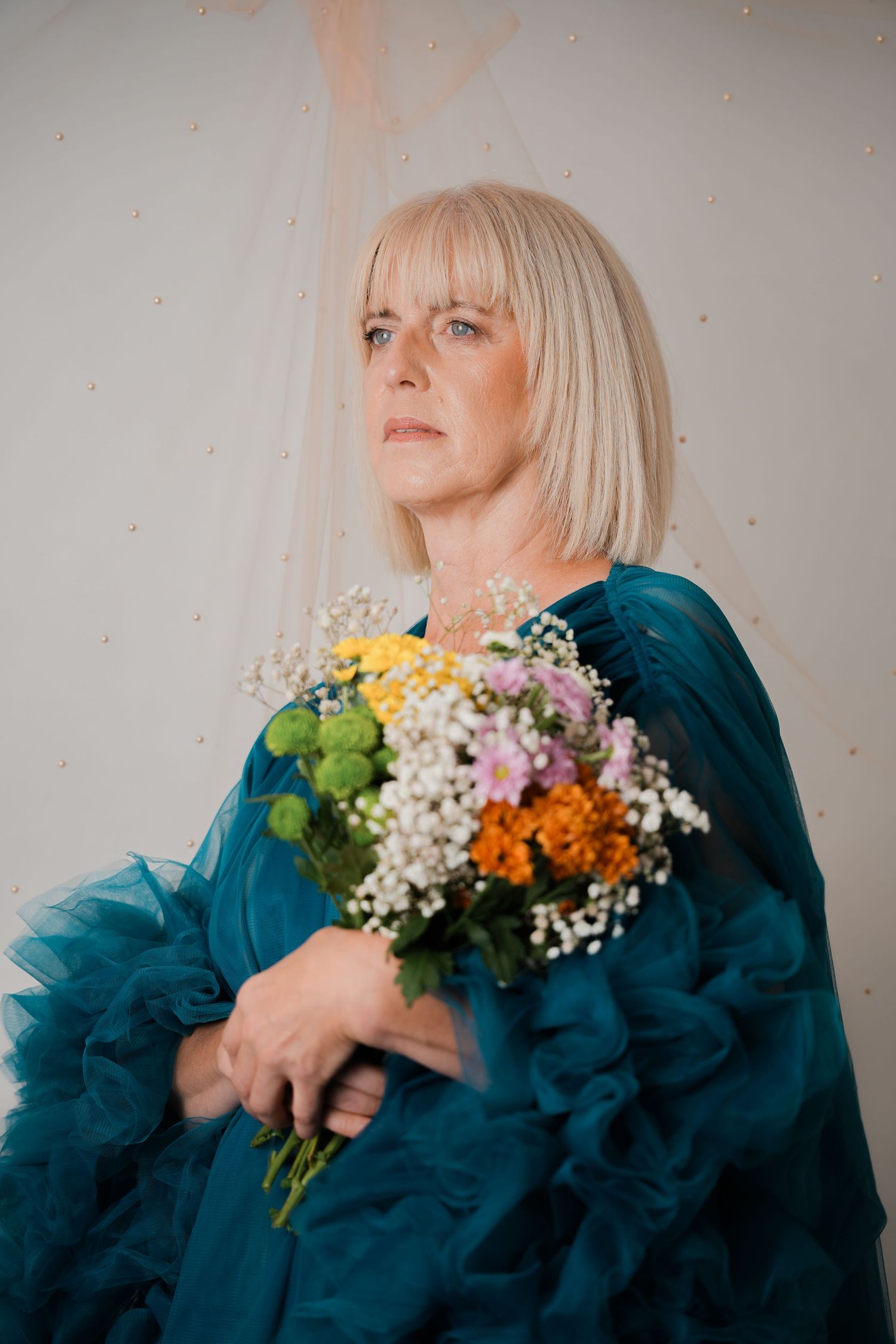 Blonde woman in teal ruffled gown holding colorful wildflower bouquet, posing thoughtfully against light background.