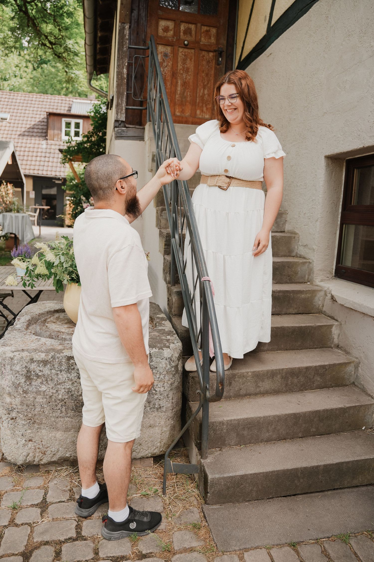 A man holds a woman's hand as she descends stone stairs outside a rustic building, both dressed in white outfits.