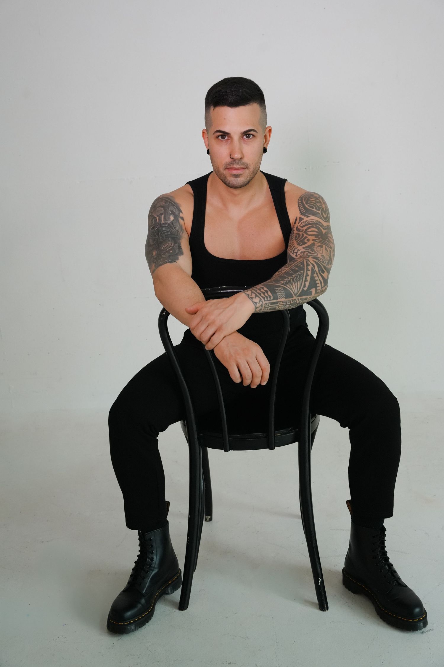 Tattooed man in black tank top and pants sitting on chair against white background.