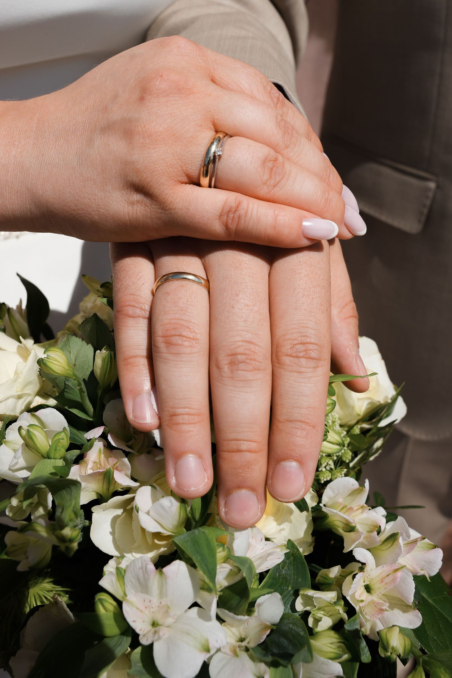 Two newlyweds display their gold wedding rings while holding hands over a bouquet of white flowers.