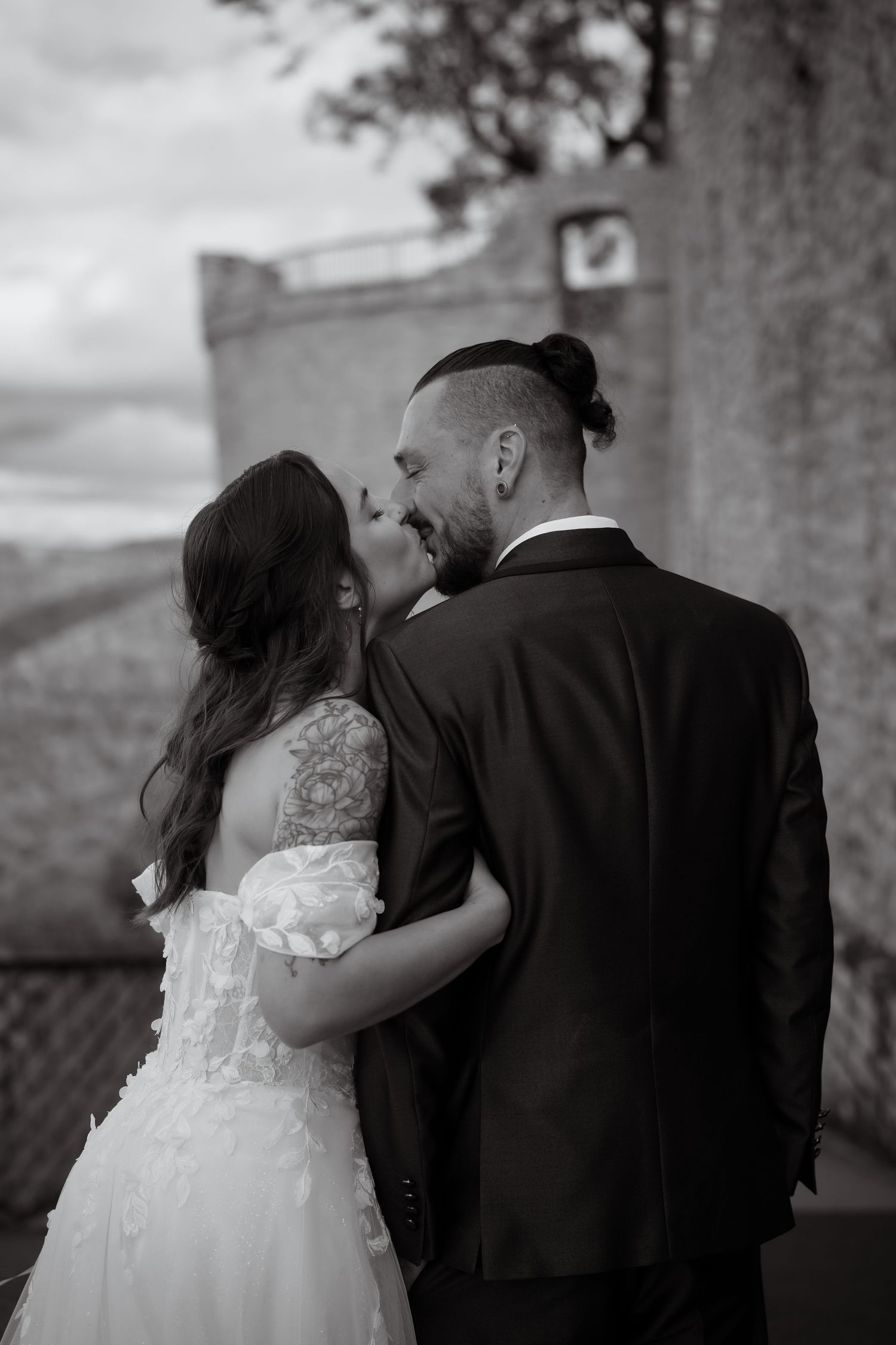 Black and white photo of a bride in a lace wedding gown kissing a groom with a top knot and dark suit near a stone building.