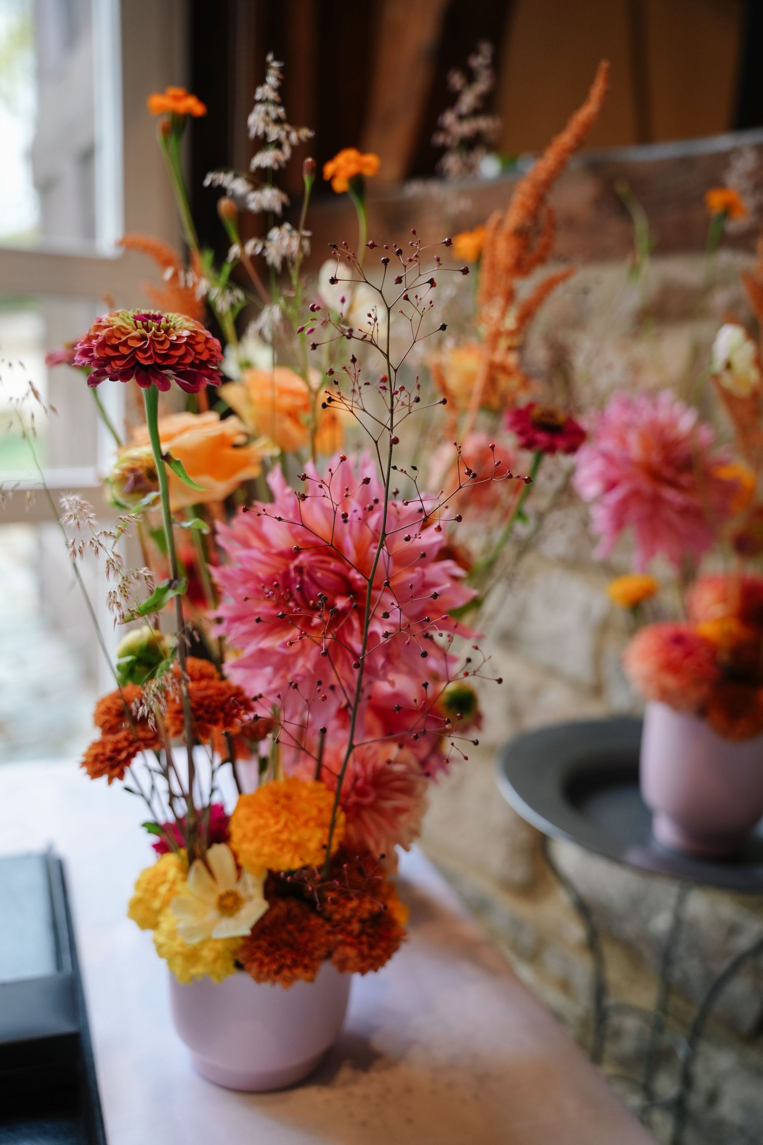 Autumn flower arrangement in a pink vase with pink dahlias, orange zinnias, yellow blooms and delicate wispy stems near a stone wall.