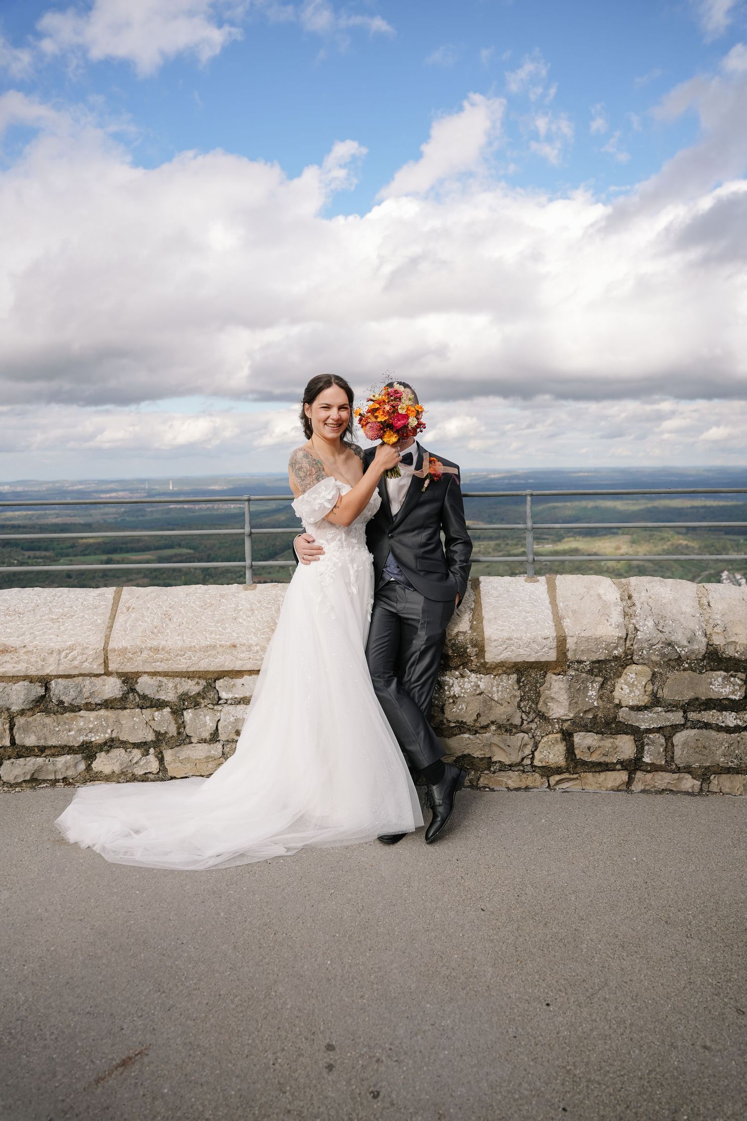Bride in a white lace gown holds an orange bouquet in front of a groom's face on a stone terrace overlooking a scenic landscape.