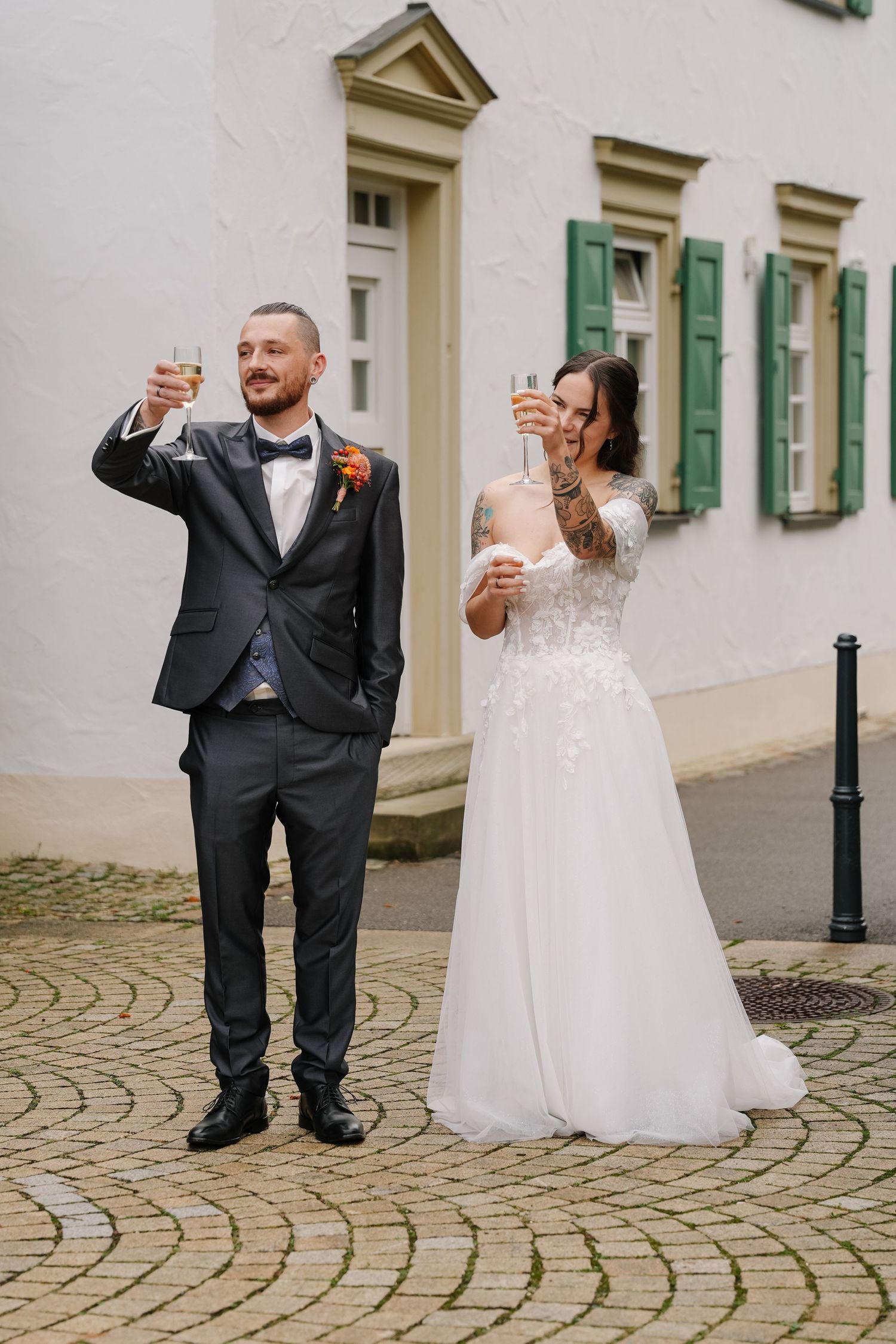 Bride and groom raise champagne glasses in a toast on a cobblestone street outside a white building with green shutters.