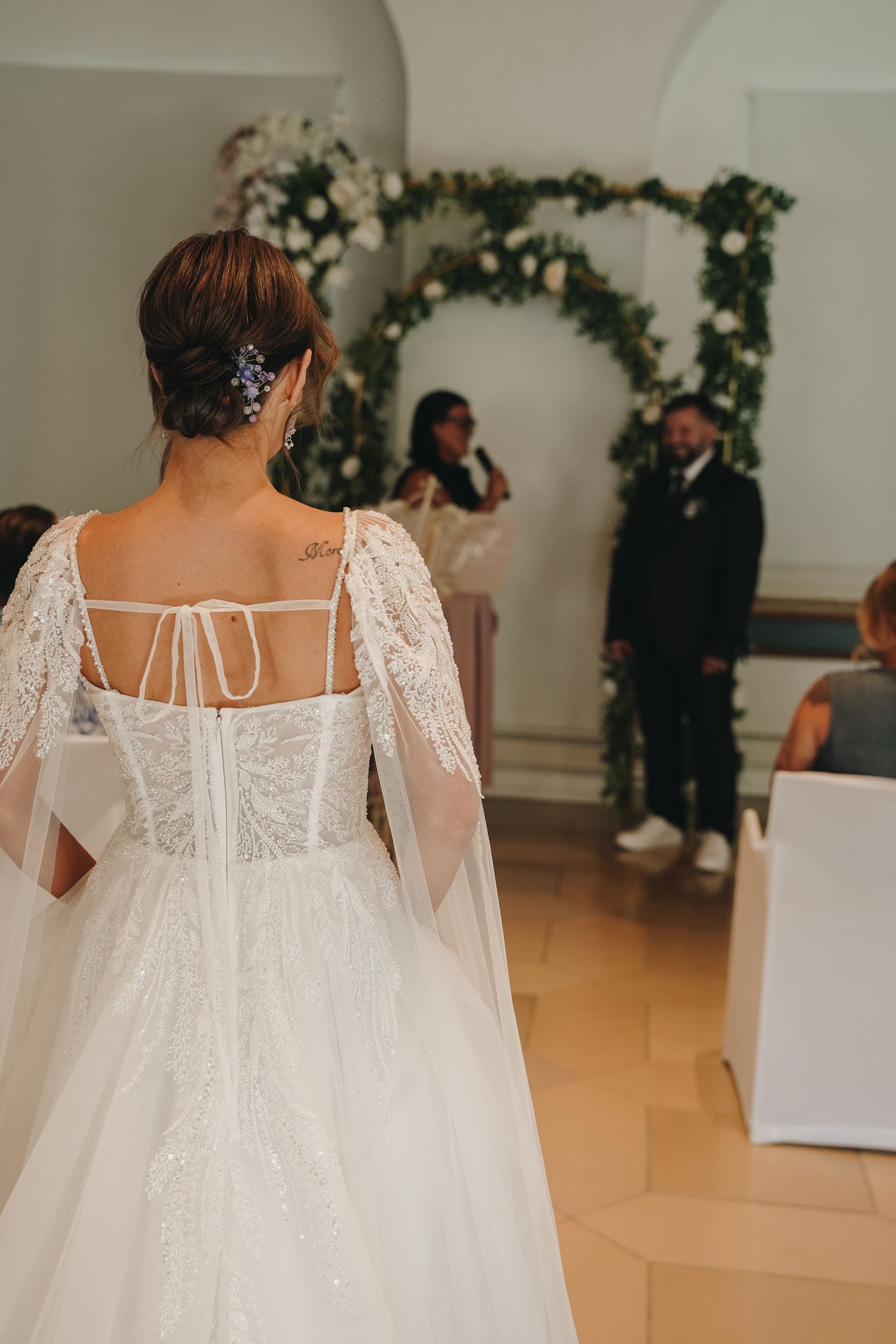 Bride in a white lace ballgown with tulle cape walks down the aisle toward groom at floral arch ceremony.
