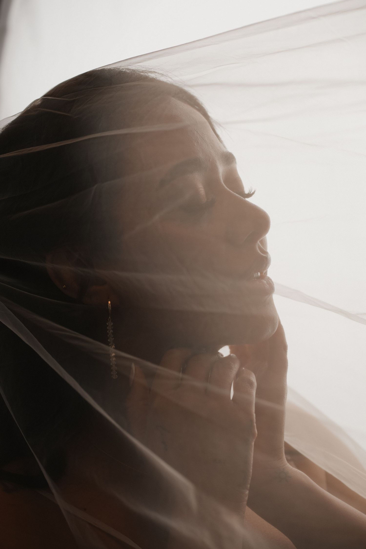 Bride draped in sheer wedding veil, soft backlit glow highlighting elegant earrings and serene expression.
