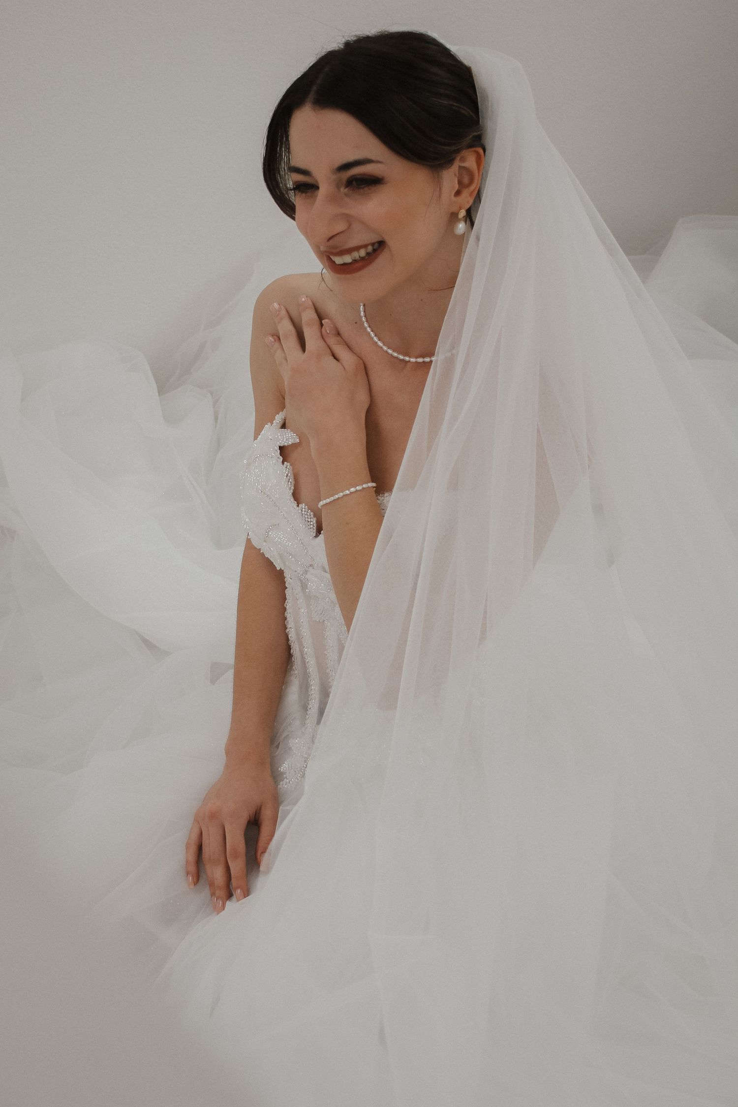 Smiling bride in white wedding gown and veil, wearing pearl necklace and bracelet, posing elegantly.