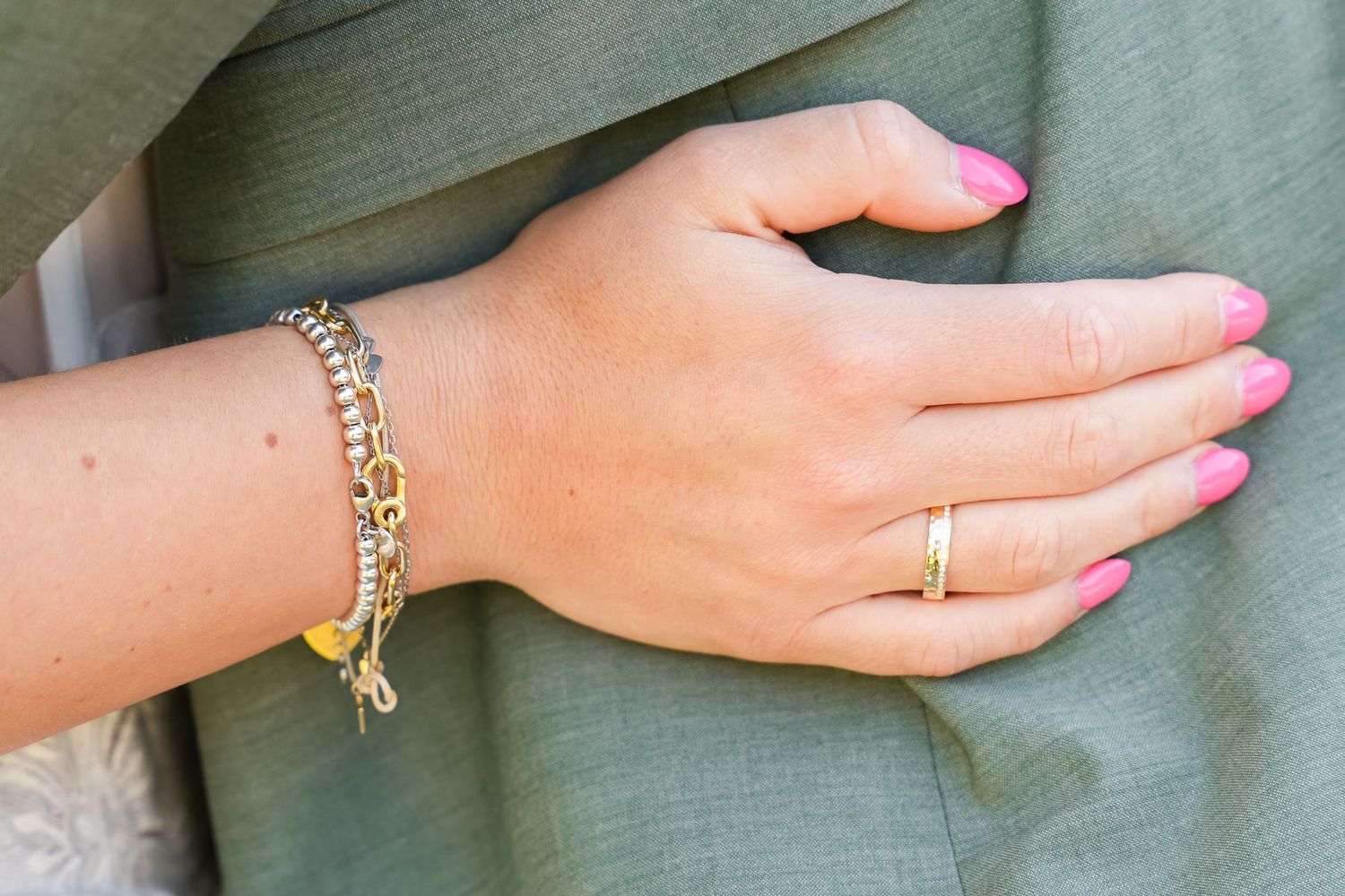 Hand with pink nails wearing gold bracelets and a gold ring against a sage green fabric background.