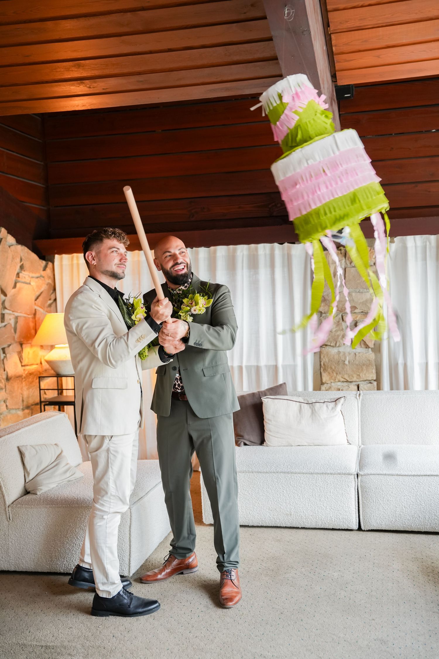 Two grooms in suits joyfully hitting a colorful tiered cake piñata with a baseball bat at their wedding.