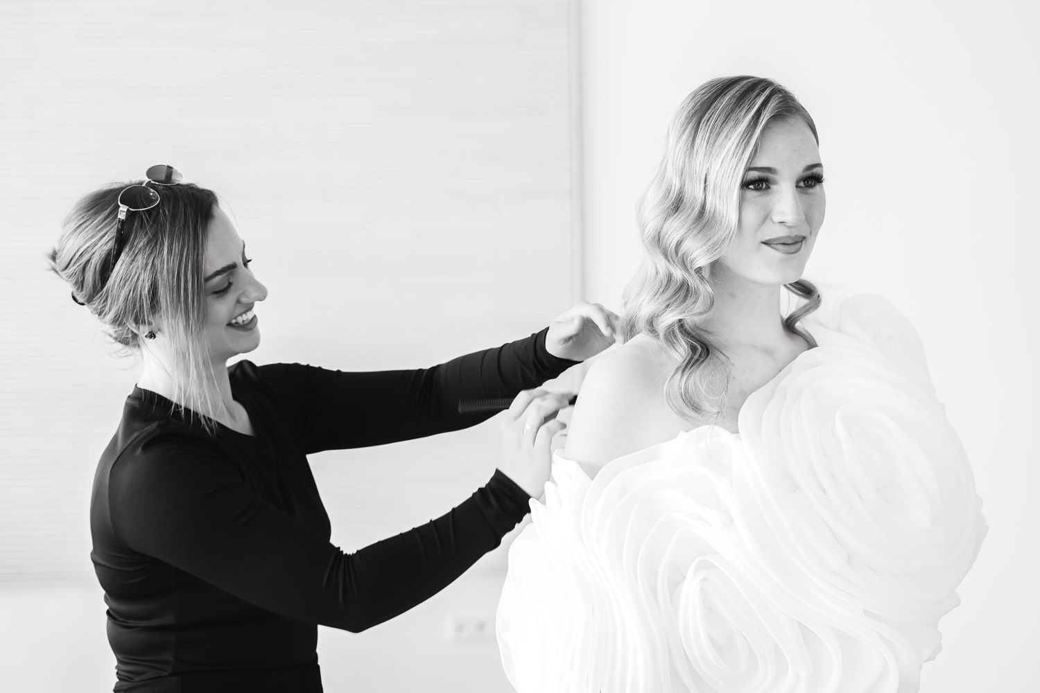 Black and white photo of a woman helping a bride in a ruffled gown with her hair on her wedding day.