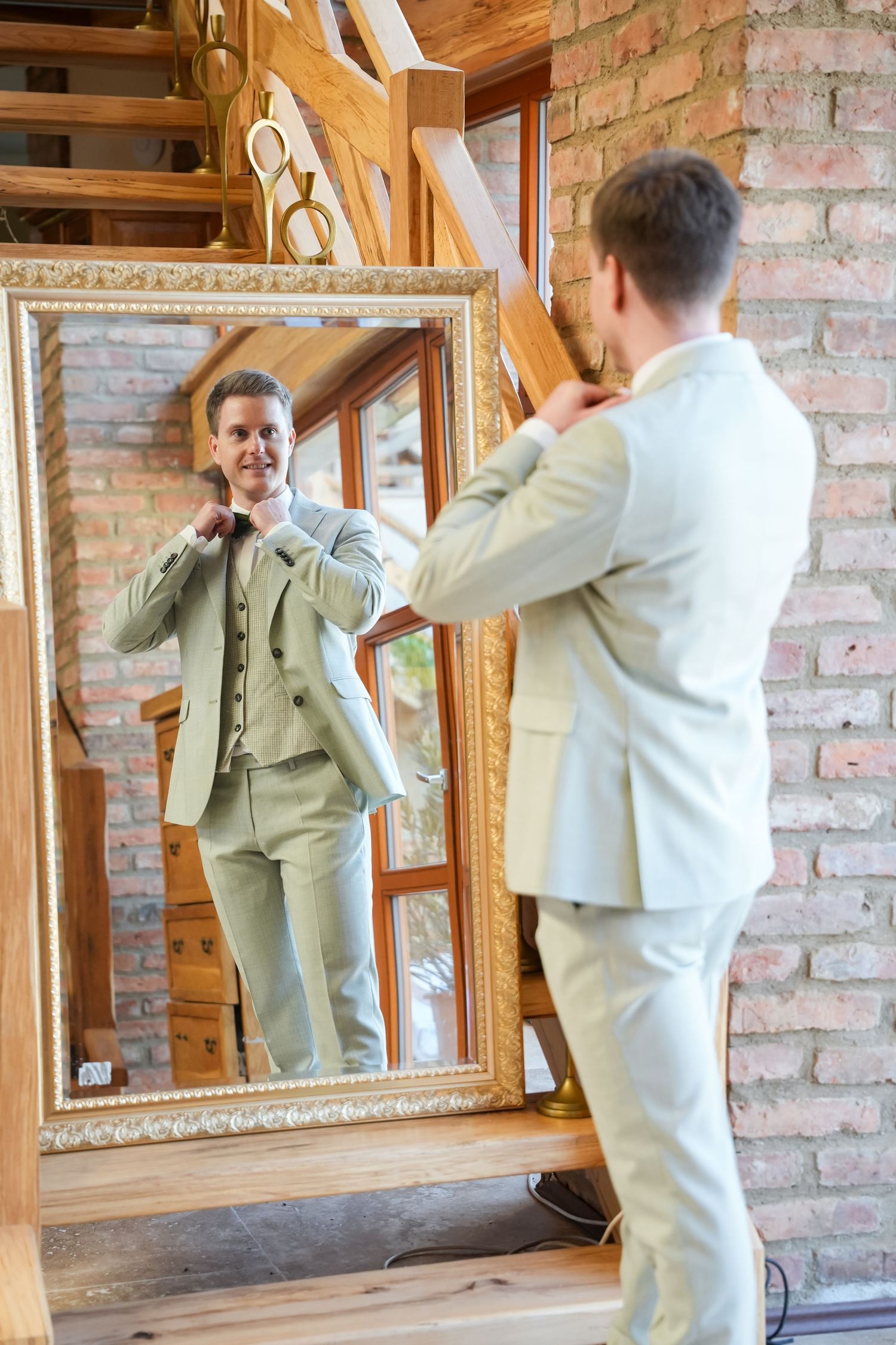A smiling groom in a light beige suit adjusts his bow tie while looking at his reflection in an ornate gold-framed mirror.
